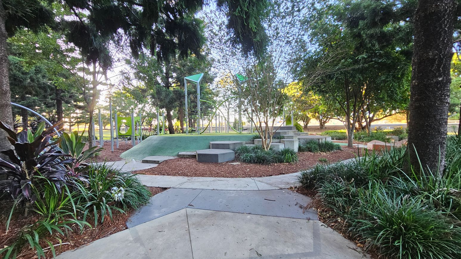 A park scene with a concrete path in the foreground leading to a play area in the middle. The play area features a green mound with geometric climbing structures and steps. Surrounding the play area are various trees and shrubs, providing shade and greenery. On the left, there are tall trees and more plants, while on the right, additional trees and a bench are visible. The background shows more trees and a hint of sunlight filtering through.
