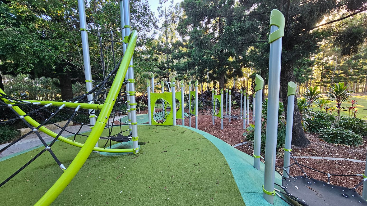 A playground with green and grey climbing structures and nets. On the left, a large netted climbing frame with metal poles. In the centre, a series of vertical poles with circular cutouts and nets. The ground is covered with green and blue safety flooring. In the background, there are tall trees and some shrubs, with sunlight filtering through the leaves.