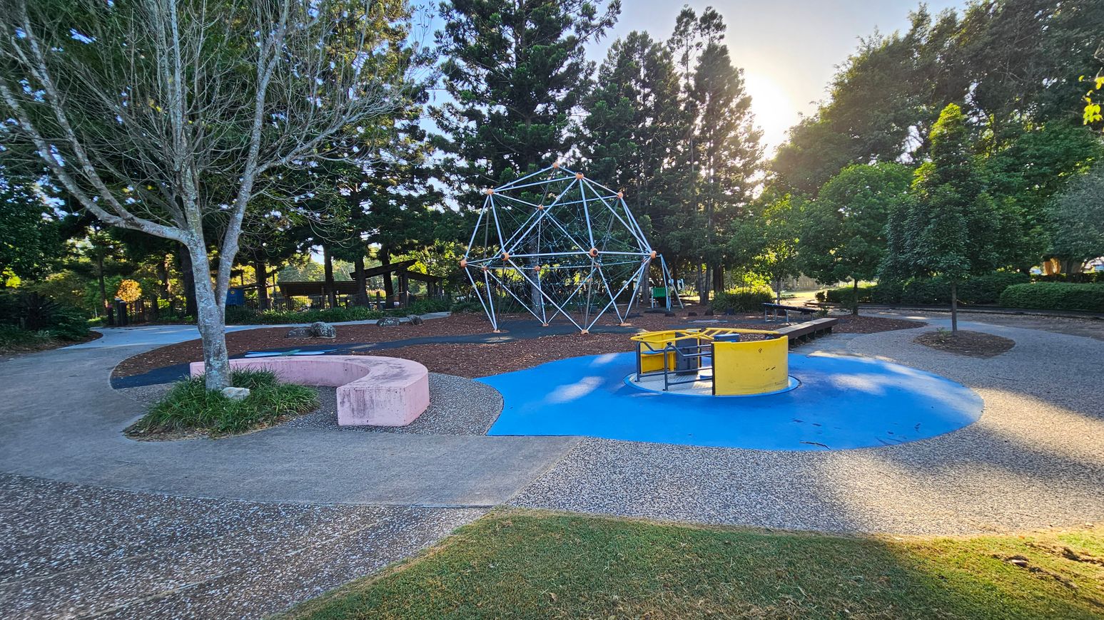 A playground with a geometric climbing structure in the centre, surrounded by tall trees. On the left, a bare tree stands next to a curved pink concrete bench. On the right, a yellow and blue roundabout sits on a blue rubber surface. The ground is a mix of grass, concrete, and pebbled paths. The background is filled with dense greenery and more trees, with sunlight filtering through.