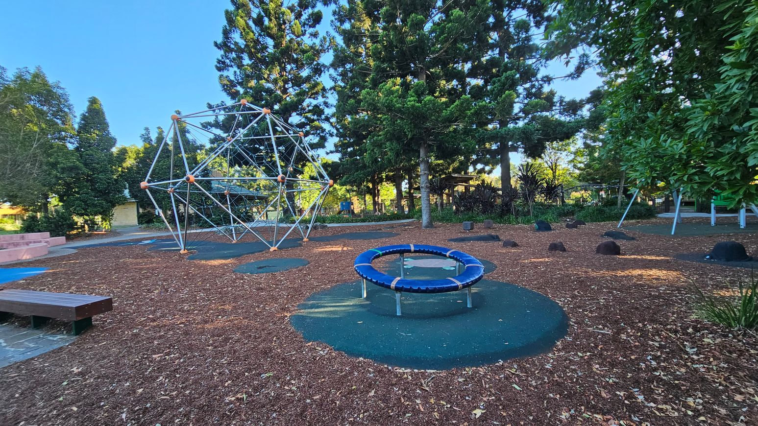 A playground with a geometric climbing structure on the left and a circular trampoline in the centre. The ground is covered with wood chips and patches of rubber matting. Tall trees surround the area, providing shade. A wooden bench is visible on the left, and there are scattered rocks on the ground. In the background, more playground equipment and dense foliage can be seen.
