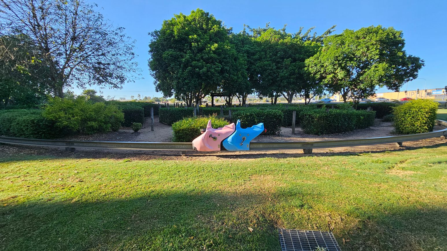 A park scene with a grassy foreground and a metal grate on the ground. In the centre, there are two animal-shaped play structures, one pink and one blue, positioned on a metal barrier. Behind them, a path is lined with neatly trimmed hedges and several large, leafy trees. In the background, there are more trees and a glimpse of a road with vehicles. The sky is clear and blue.