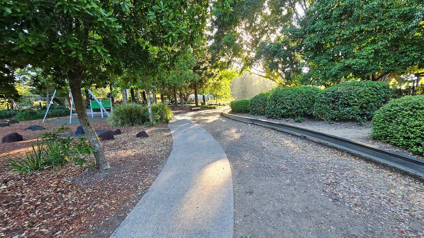 A park scene with a winding concrete path in the centre, surrounded by trees and bushes. On the left, there's a playground area with a swing set and a green bench. The ground is covered with mulch and scattered leaves. On the right, a narrow track runs parallel to the path, bordered by neatly trimmed bushes. Sunlight filters through the trees, creating dappled light on the ground.