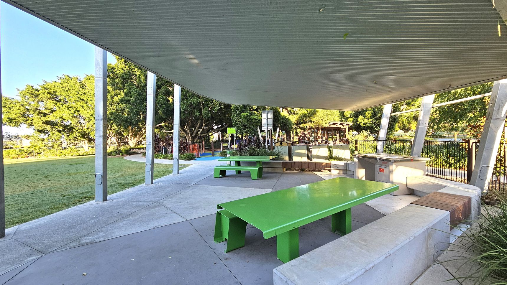 A covered outdoor area with a corrugated metal roof supported by metal poles. In the foreground, there is a bright green picnic table with attached benches on a concrete floor. In the background, another similar table is visible, along with a barbecue station and a rubbish bin. The area is surrounded by a grassy lawn and trees, with a playground partially visible in the distance. The scene is well-lit, suggesting daytime.