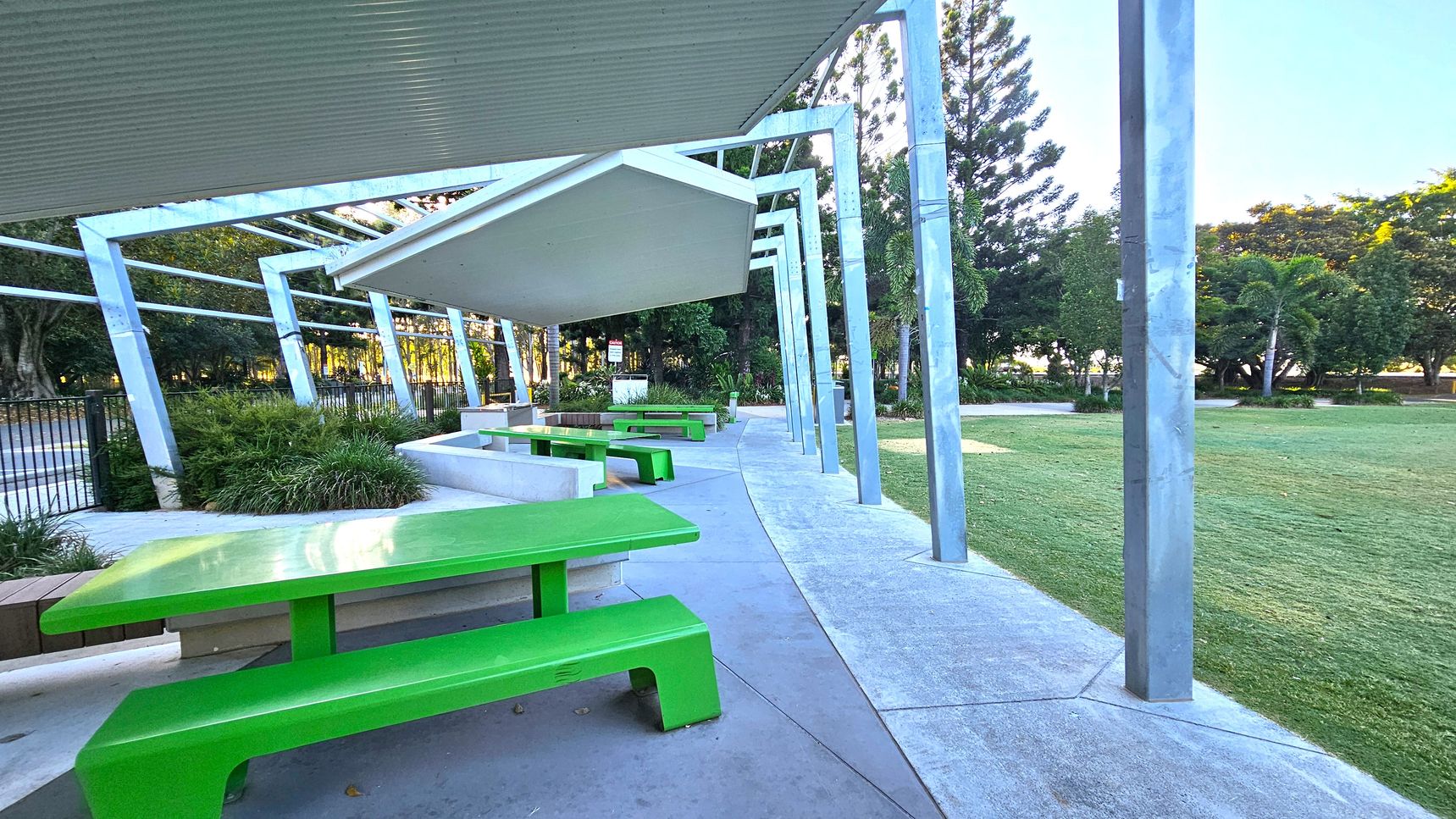 A covered outdoor area with a series of green picnic tables and benches on a concrete floor. The structure has a metal roof supported by angled metal beams. To the left, there are plants and a fence, while to the right, a grassy lawn extends with trees in the background. The ceiling is corrugated metal, and the area is open to the surrounding park.