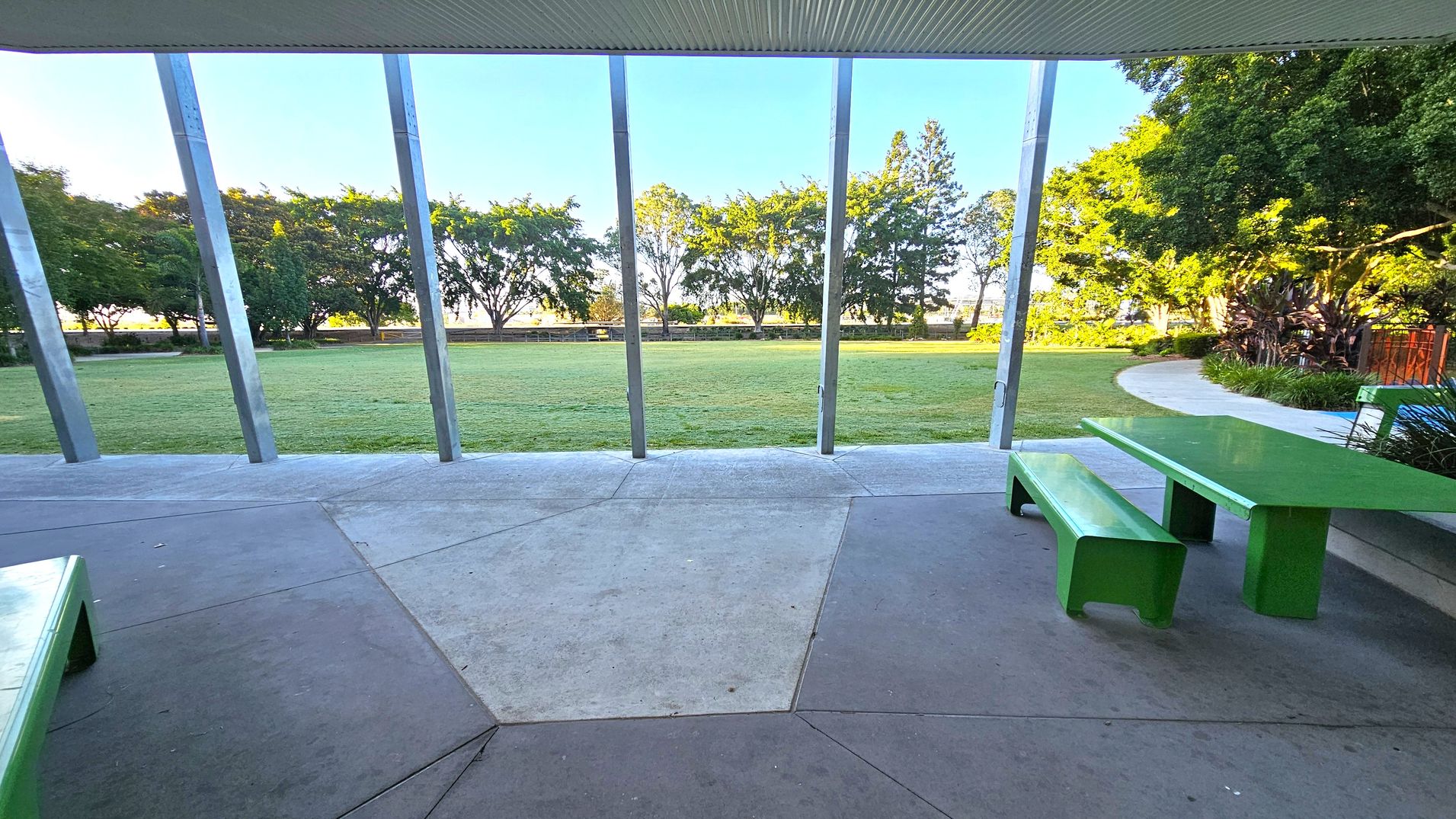 A covered outdoor area with a green picnic table and bench on the right. The floor is made of concrete with geometric patterns. In the background, a grassy field is bordered by a row of trees. Metal beams support the structure, and a pathway curves around the right side, lined with plants. The ceiling is corrugated.