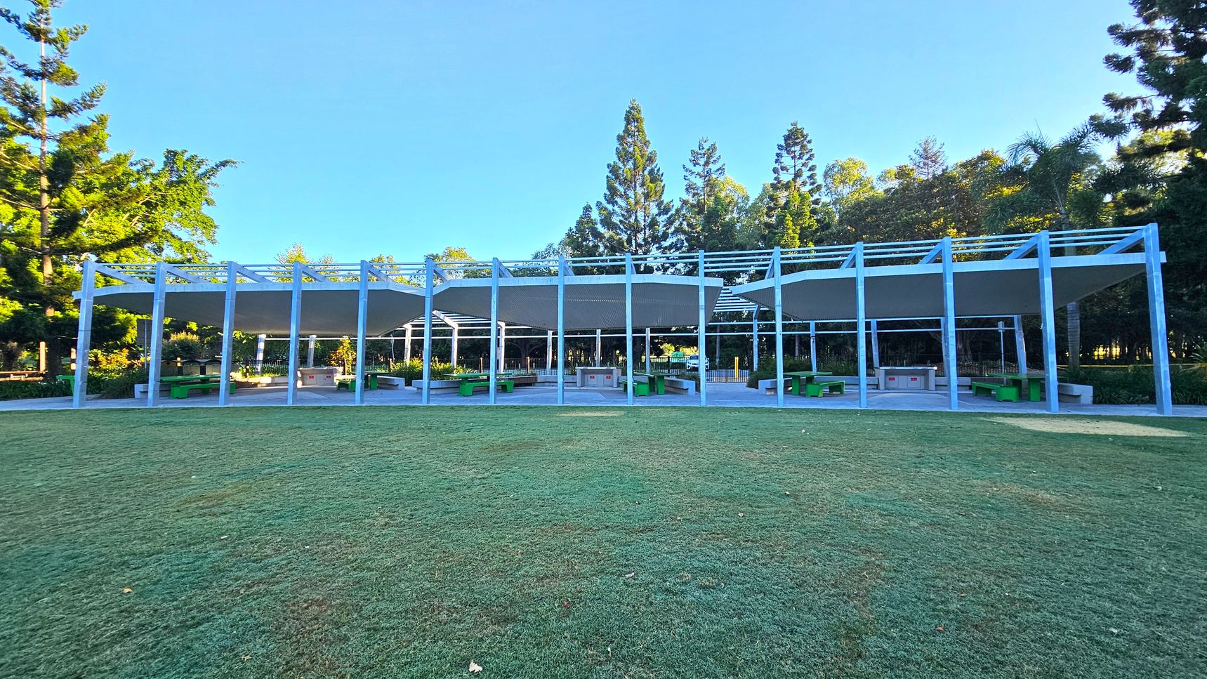 A grassy area in the foreground leads to a modern outdoor pavilion with a series of blue metal frames and white canopies. Beneath the canopies are green benches and tables, with a barbecue area in the centre. Tall trees with dense foliage form the background, providing a natural backdrop to the structure. The scene is well-lit, suggesting daylight.