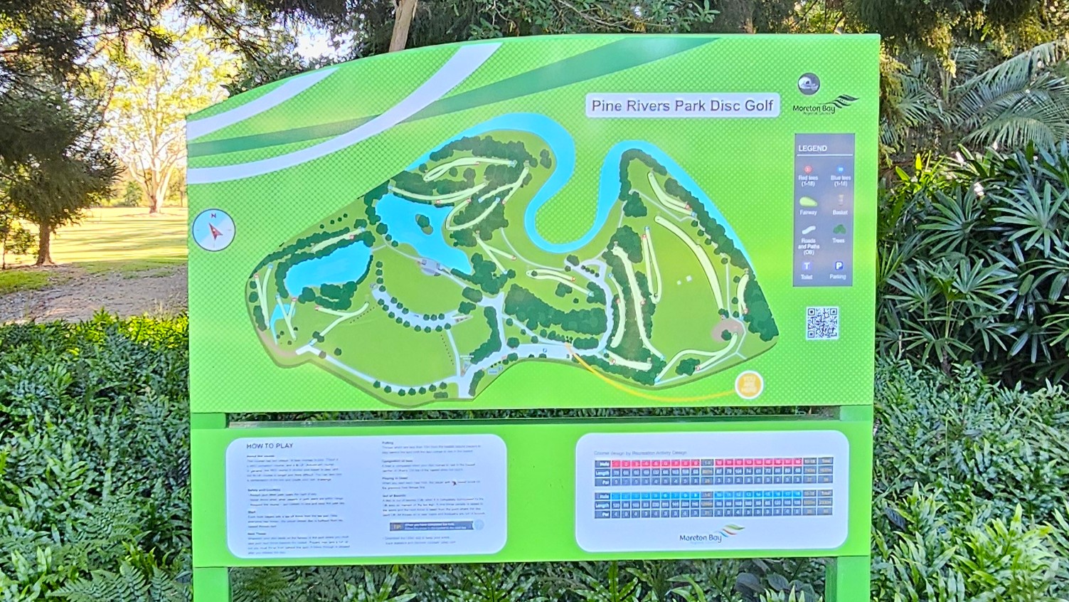 A green information board displaying a map of Pine Rivers Park Disc Golf course. The map features pathways, water bodies, and various course details. On the right, there's a legend with symbols and a QR code. Below the map, there are instructions and a scorecard. The board is surrounded by lush greenery, with trees and bushes in the background.