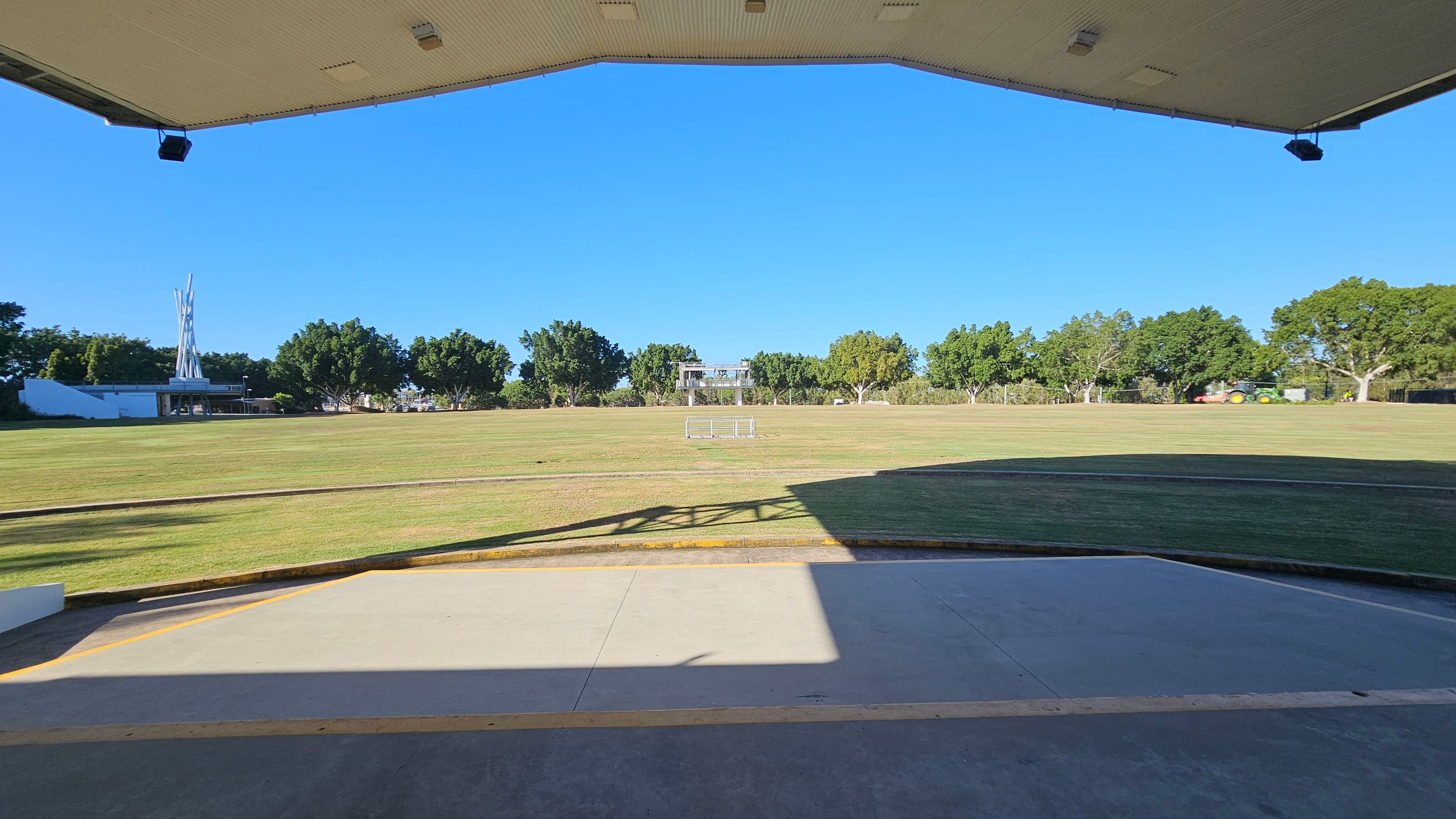 A large open grassy field is visible, bordered by trees in the background. In the centre, there is a small structure or platform. To the left, a tall, white, abstract sculpture stands near a building. The foreground shows a concrete area with shadows cast from a structure above, which is partially visible at the top of the image. The sky is clear and blue.