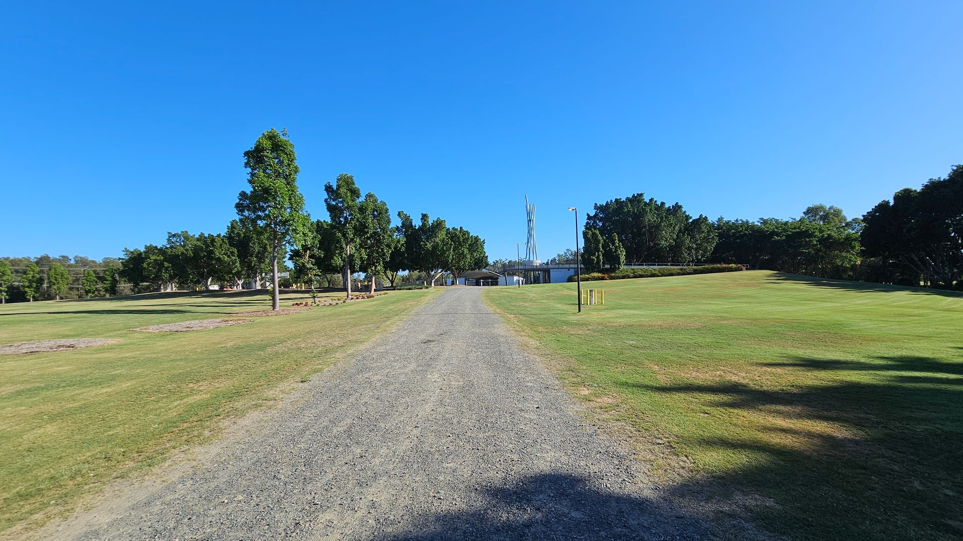 A gravel path runs through the centre of a grassy area, leading towards a structure in the distance. Trees line both sides of the path, with more dense foliage on the right. On the left, the grass appears slightly patchy. In the background, a tall, slender structure is visible, surrounded by more trees and a few buildings. The sky is clear and blue.