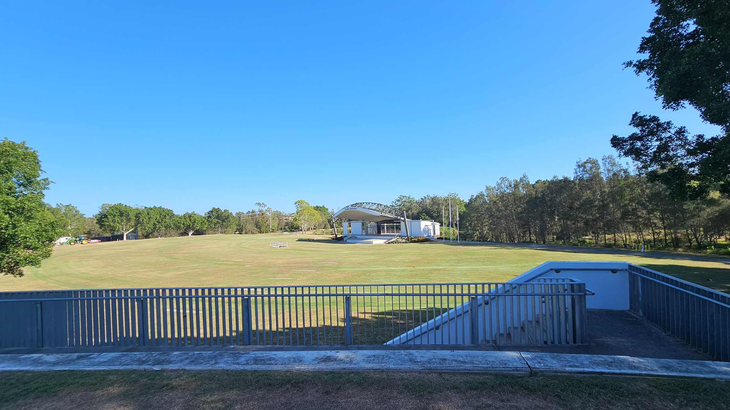 A grassy field with a stage structure in the centre, featuring a curved roof and white walls. Trees line the background, providing a natural border. On the left, a tractor is visible near a small building. In the foreground, a concrete path and a railing lead down to a lower level, with a staircase descending on the right. Trees frame the scene on both sides.