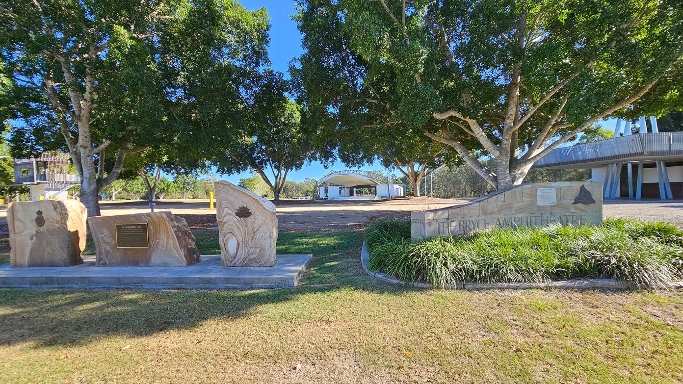 In the foreground, there are three large stone slabs with engravings and a plaque. To the right, a sign reads "The Bryce Amphitheatre" with surrounding greenery. In the background, a large amphitheatre structure is visible, partially obscured by two large trees with dense foliage. The ground is covered with grass, and there is a paved area near the stones.