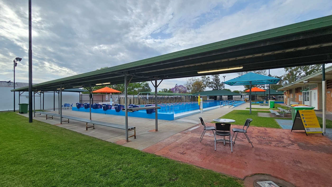 Outdoor swimming pool area with a long, rectangular pool in the centre. On the left, there is a covered seating area with chairs. The right side features a grassy area with a bench and green bins. Blue and orange shade structures are positioned over the poolside. A yellow sign with safety instructions is in the foreground. The background includes trees and a fence, with a cloudy sky above. The ground is a mix of concrete and grass.