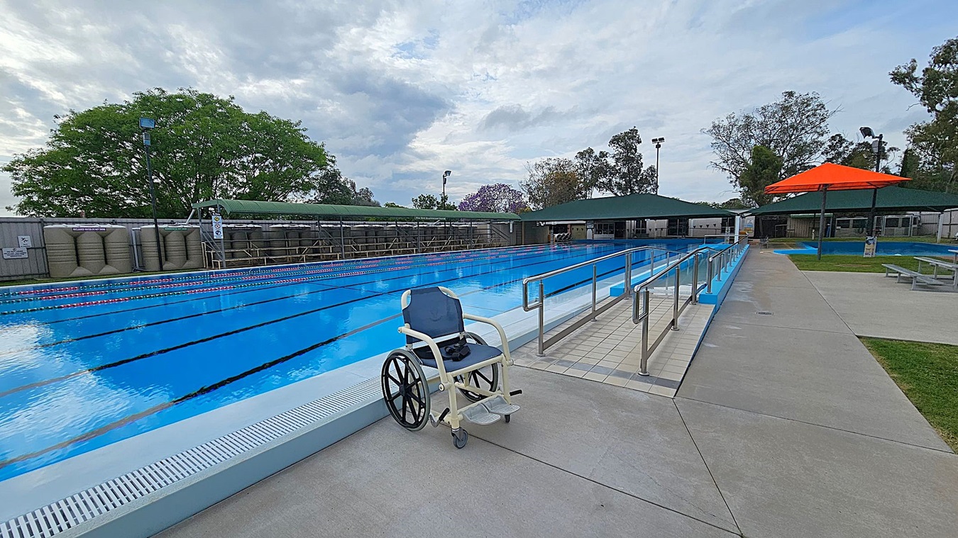 A swimming pool with clear blue water and lane dividers is on the left. A wheelchair is positioned near the pool's edge. On the right, a paved walkway with metal railings leads to the pool. In the background, there are green-roofed structures and a large orange umbrella over a seating area. Trees with green and purple foliage are visible beyond the pool area. The ground is concrete, and the area appears to be an outdoor facility.