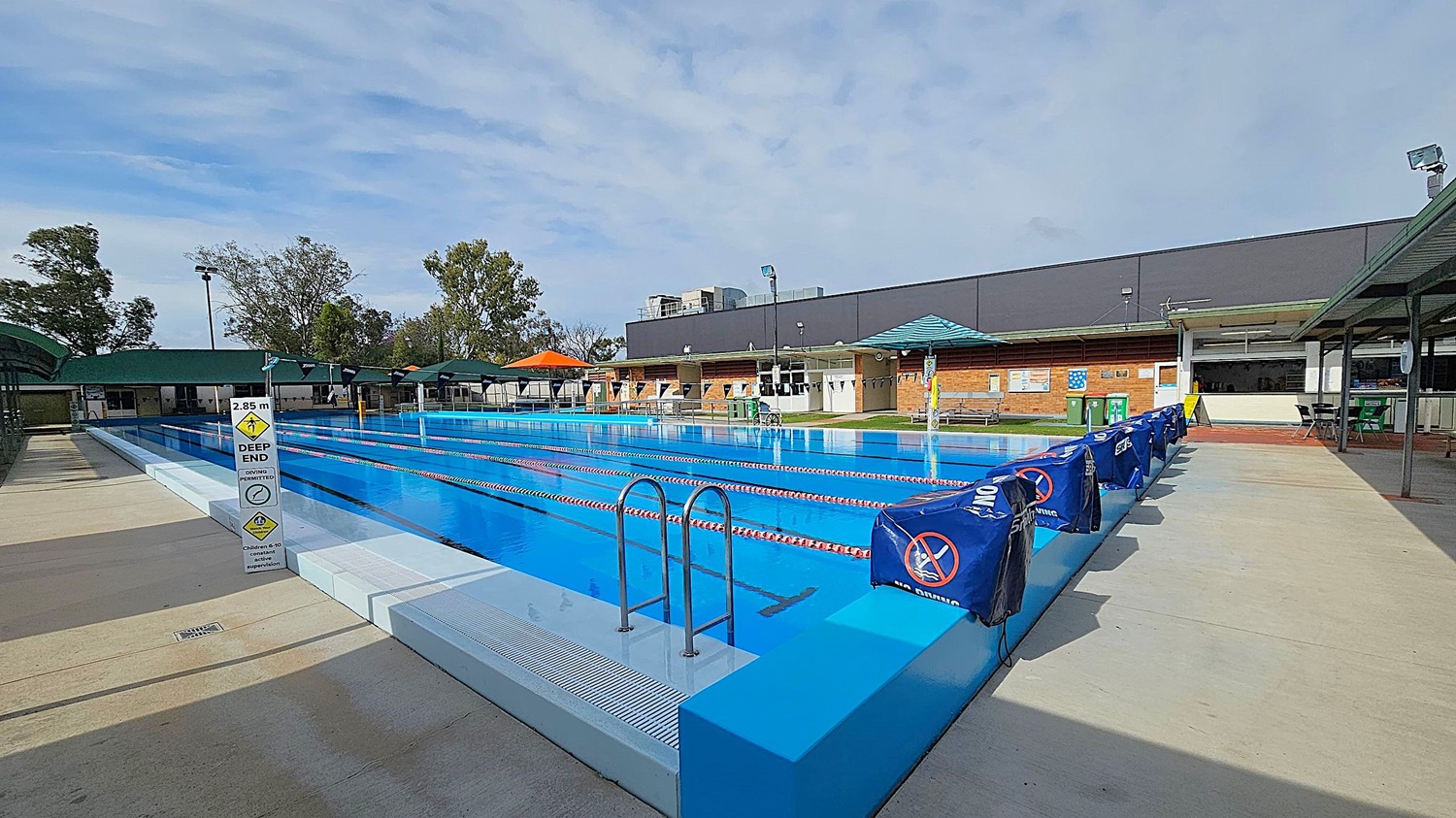 Outdoor swimming pool with clear blue water and lane dividers. A sign indicates a depth of 2.85 metres at the deep end. On the left, a covered seating area with green roofs. On the right, a row of blue covers with no diving signs. In the background, a brick building with a green roof and several trees. The pool deck is concrete.