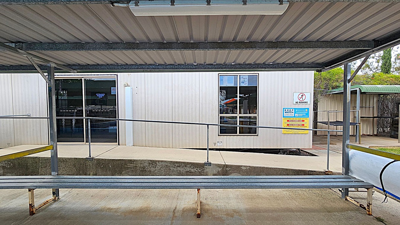 A metal shelter with a corrugated roof covers a walkway leading to a building with a glass door and a window. The building is light-coloured with a sign next to the window displaying safety instructions and a "No Running" warning. The ground is concrete, and there is a metal railing along the walkway. In the background, there is greenery and another structure with a green roof.