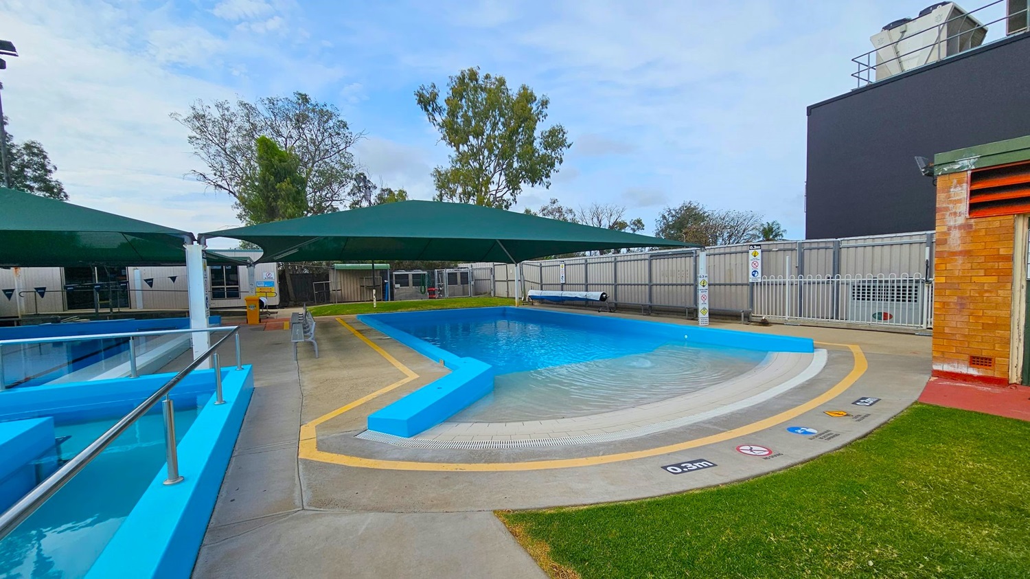 A small outdoor swimming pool with a bright blue border is in the centre, surrounded by a concrete deck with safety signs. Green shade sails cover the pool area. On the left, there is a metal railing and a smaller pool section. The right side features a brick building and a grassy patch. In the background, there are trees and a tall, dark structure. A fence encloses the area.