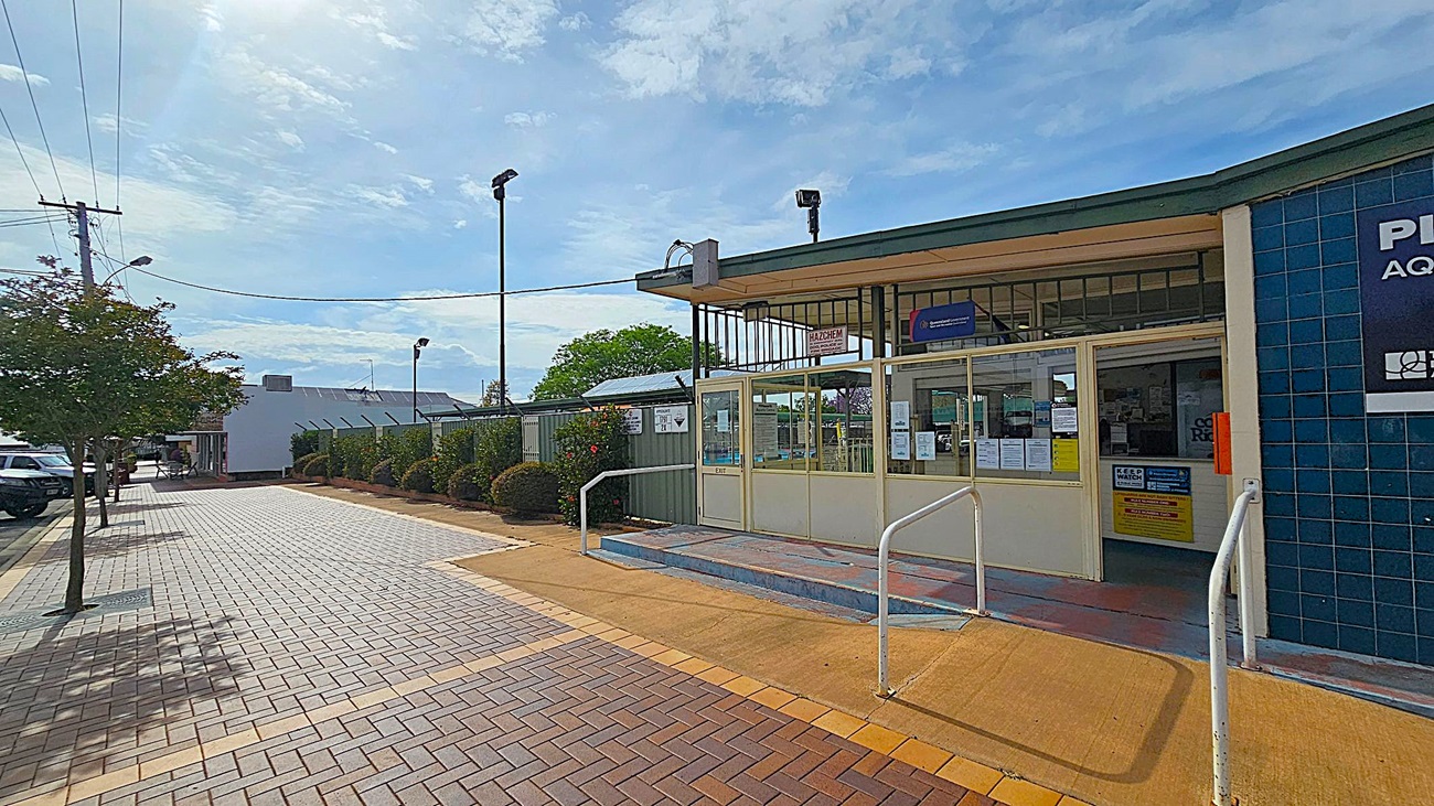 A paved walkway leads to a building entrance on the right, featuring glass doors and a blue-tiled wall with signage. The entrance has a ramp with white railings. On the left, a row of small trees lines the walkway, with parked cars visible further down. The background includes a fence with bushes and a building with solar panels. Overhead, power lines stretch across the scene.