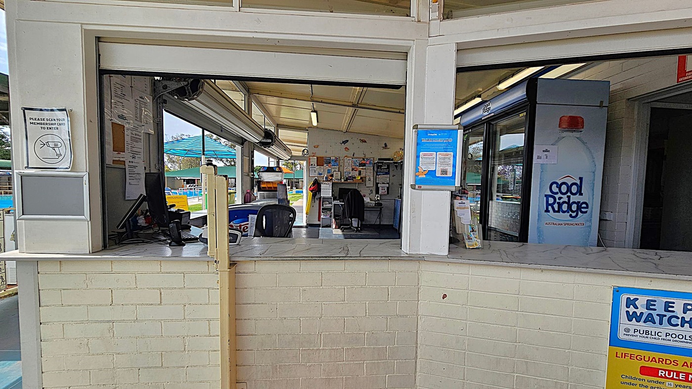 A reception area with a marble counter and a light green brick wall. On the left, there's a sign instructing to scan a membership card. Behind the counter, there are two chairs, a computer, and a wall covered with notices and posters. On the right, a large fridge displays a "Cool Ridge" water advertisement. The ceiling is slanted with fluorescent lights, and outside, a blue shade structure is visible.