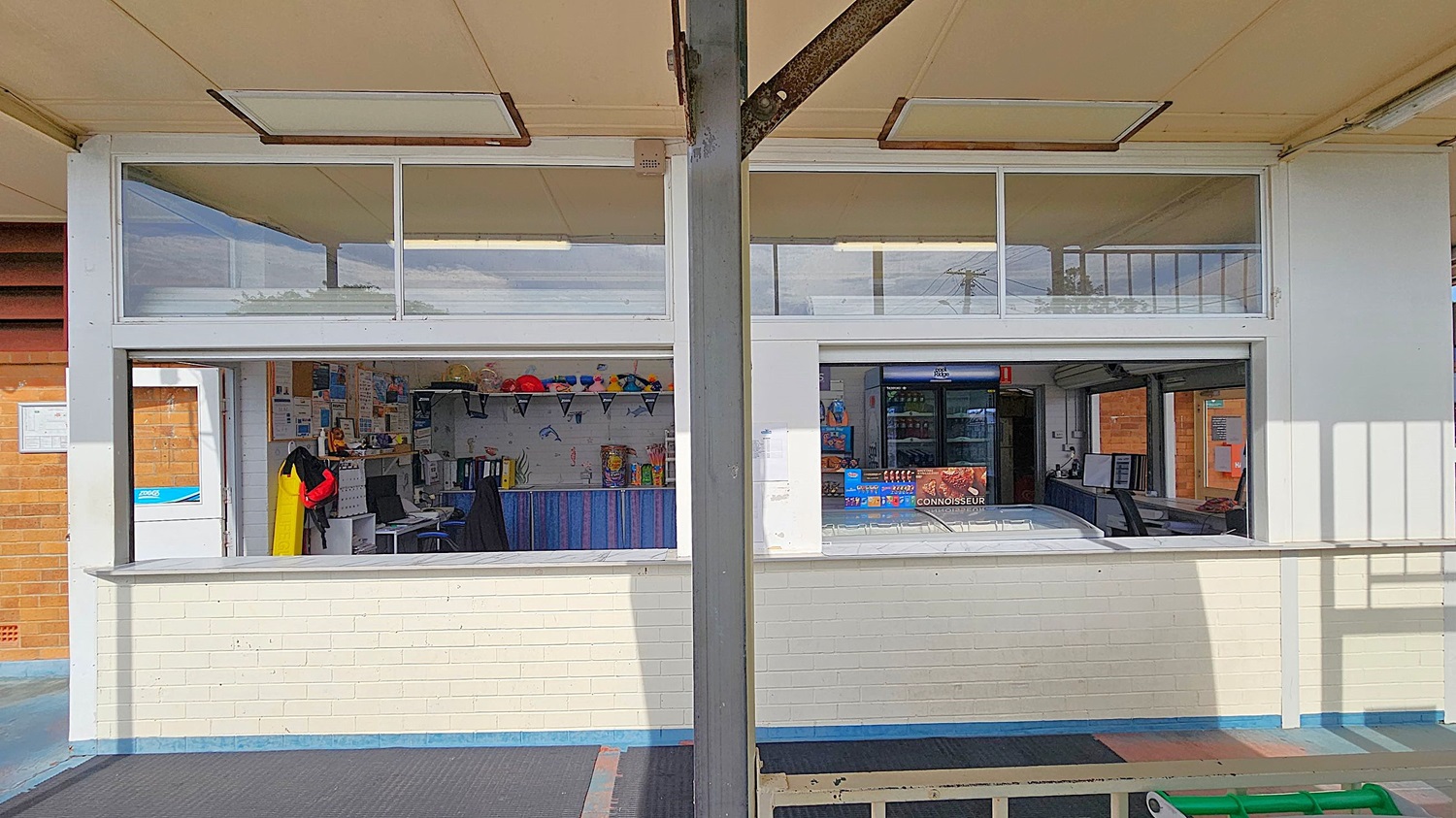 A kiosk with two service windows divided by a metal pillar. On the left, the interior has shelves with colourful items and a desk with a computer. A yellow and red jacket hangs nearby. On the right, there's a display of ice cream products and a fridge. The ceiling is white with fluorescent lights, and the floor is a mix of blue and black mats. The walls are white brick.