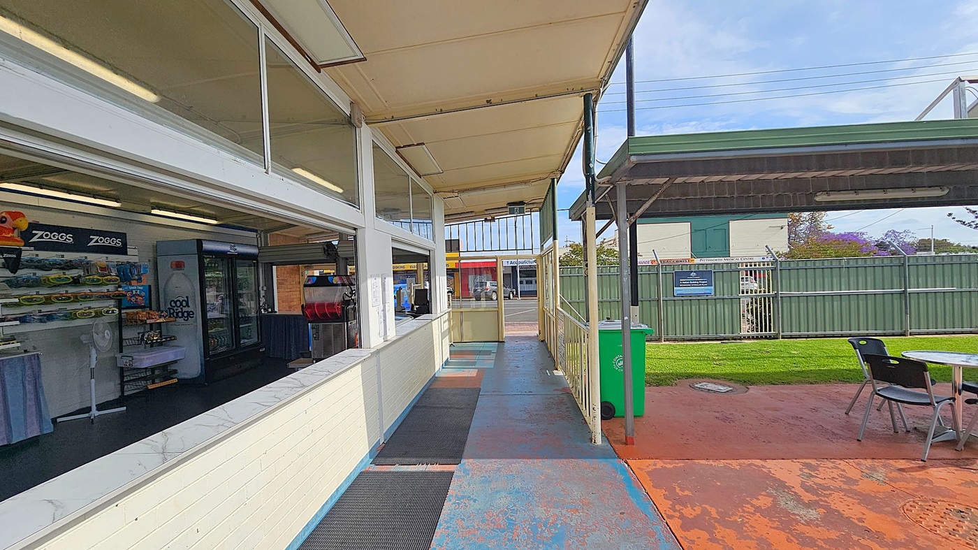 A covered walkway with a shop on the left, displaying shelves of products and a fridge with a "Cool Ridge" label. The floor is painted blue with black mats. On the right, there's a green bin and a seating area with a table and chairs on a reddish-brown concrete surface. A green metal fence and a building are visible in the background. The ceiling is beige with fluorescent lights.