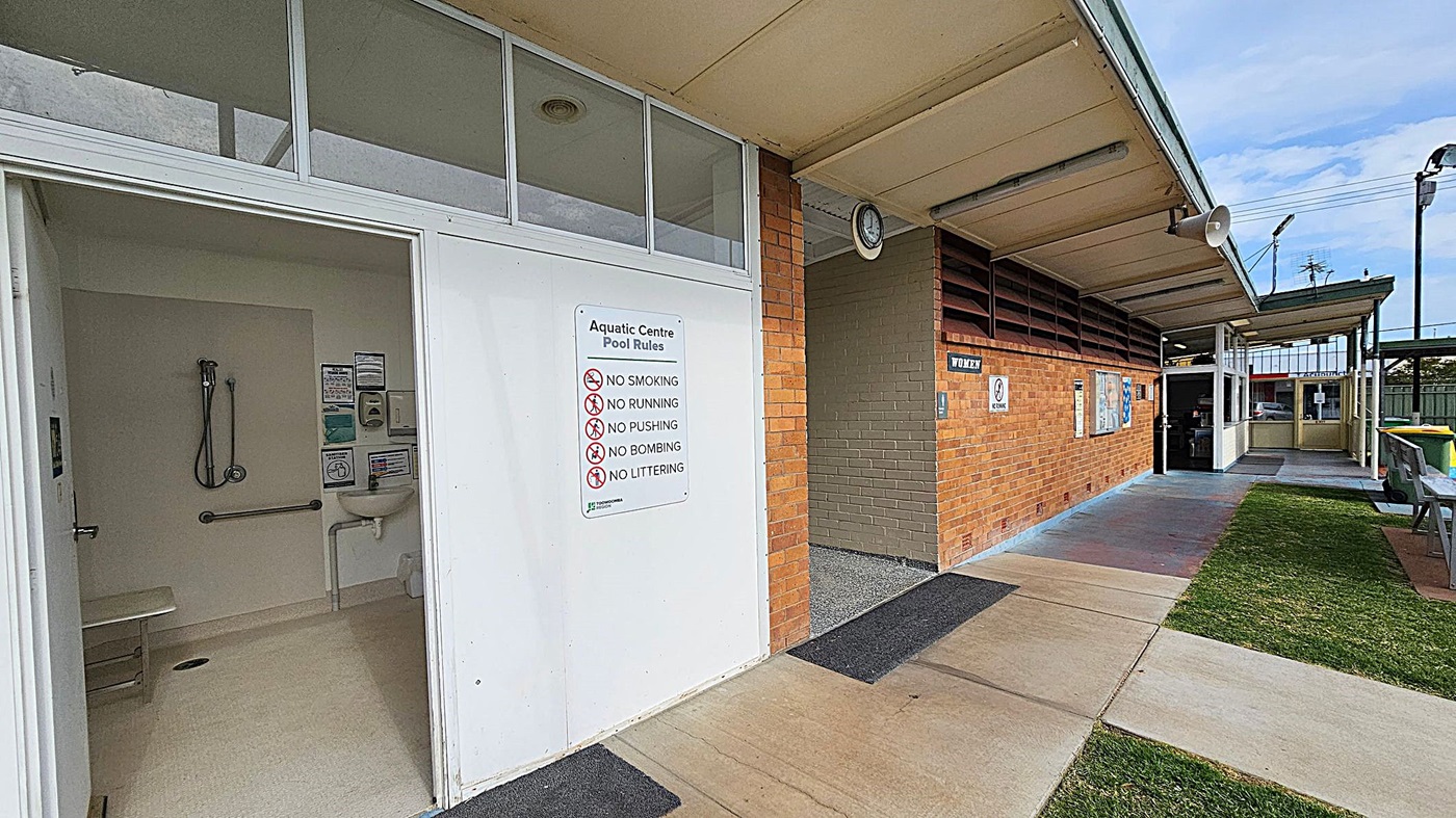 On the left, there's an open door leading to a shower room with a showerhead, grab bar, and sink. A sign on the door lists pool rules, including no smoking and no running. The middle features a brick wall with a "Women" sign and a clock above. On the right, there's a covered walkway with a bench and a green bin. The floor is a mix of concrete and grass, and the ceiling is beige with fluorescent lights.