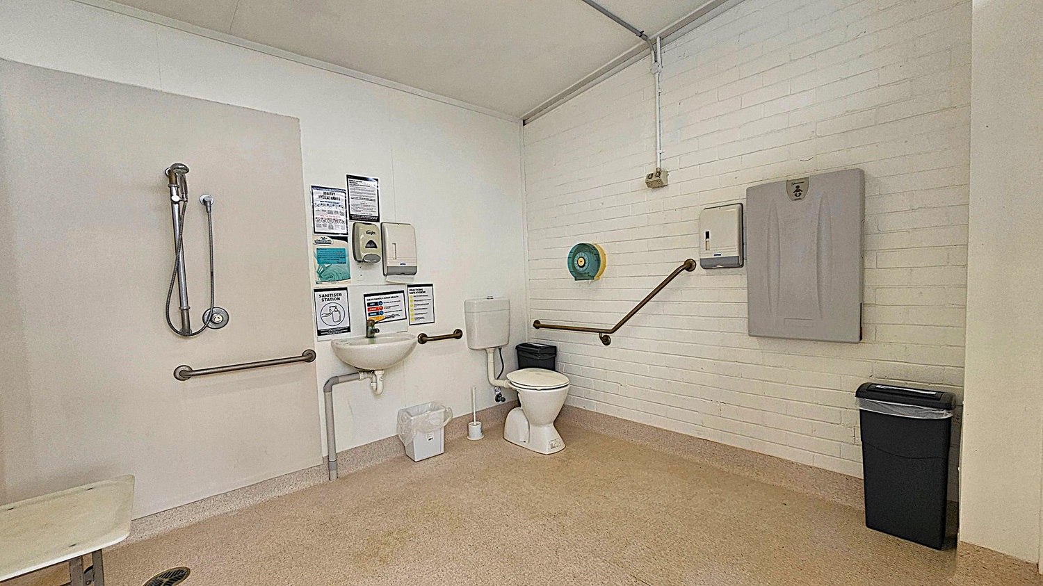 A spacious, accessible bathroom with a beige floor and white brick walls. On the left, there's a shower with a handheld nozzle and a grab bar. In the centre, a small sink with a soap dispenser and sanitiser station above it. To the right, a toilet with grab bars on both sides, a toilet paper dispenser, and a nappy changing station. A black bin is in the far right corner. The ceiling is plain white with visible piping.