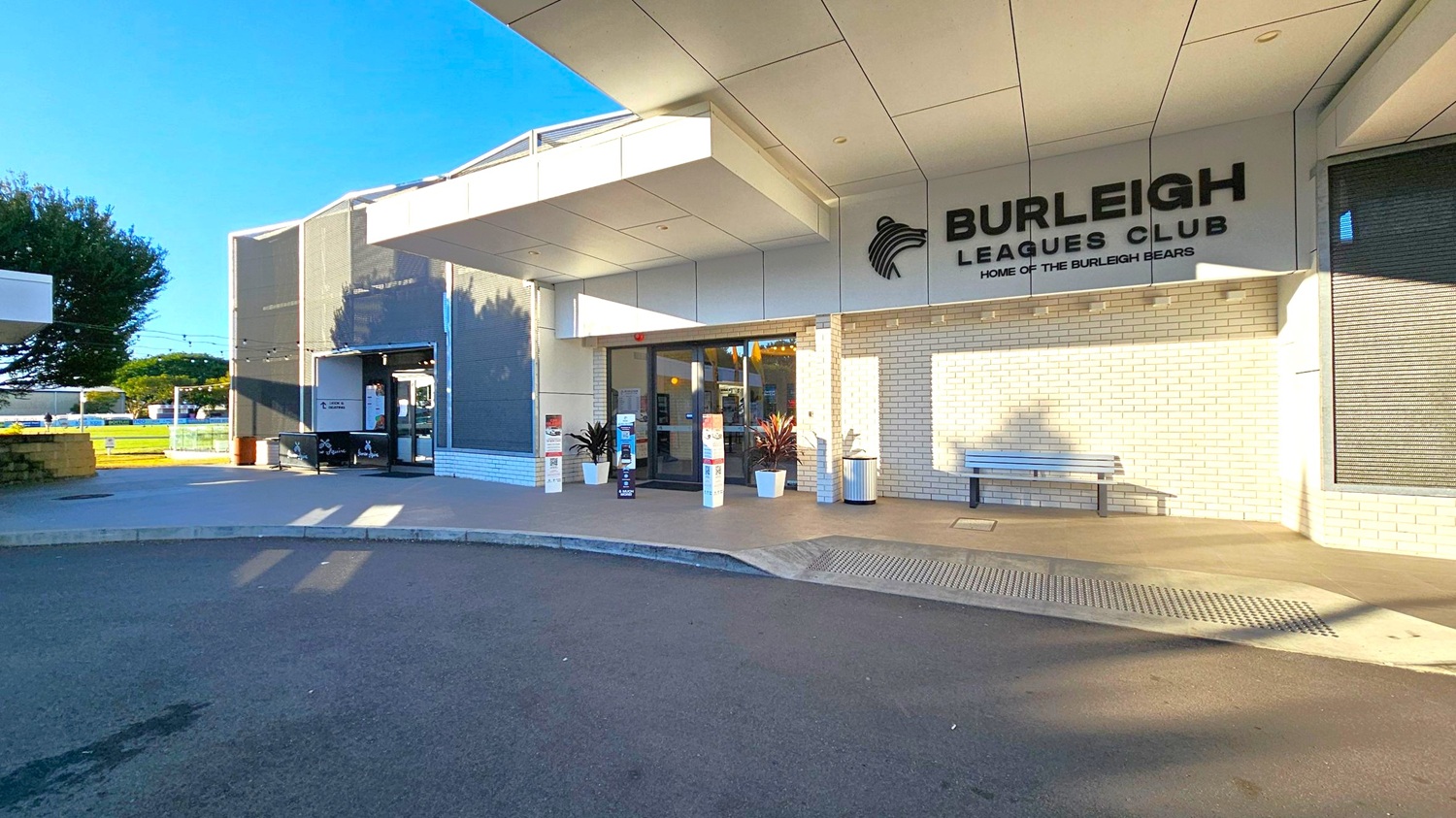 Entrance to a building with a sign reading "Burleigh Leagues Club" and "Home of the Burleigh Bears" on the right. The facade is made of light-coloured bricks. A bench and potted plants are near the entrance. The left side shows a tree and a small outdoor area with string lights. The ground is paved, and the ceiling has a modern, geometric design.