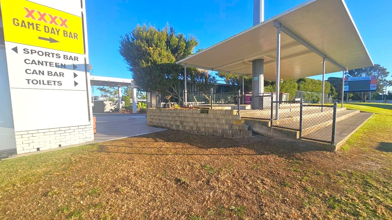 A sign on the left reads "XXXX Game Day Bar" with directions to a sports bar, canteen, can bar, and toilets. The foreground has a grassy area leading to a paved path. On the right, a covered seating area with a metal fence and a scoreboard in the background. The middle ground features a low brick wall with plants and trees providing shade. The structure has a flat roof supported by metal poles.