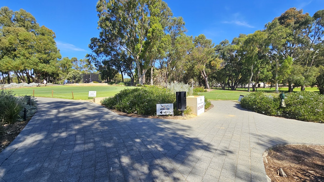 The concrete brick pathway divides; both lead to a vast green and grassy area with tall trees and shrubs. In the middle is a mini landscape with directional signages, a golf hole sponsor, and a garden lamp. The path on the left elevates towards the entrance of the building. A wooden post stands on the right along with a banner. The path on the right descends to the golf area.