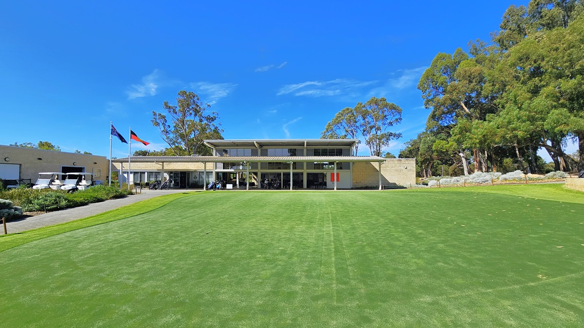 On the right, there is an expansive green space featuring well-maintained grass and synthetic turf. To the left, an ascending path leads to the building. In the middle stands a two-story cream building. Two flags are positioned on the right, accompanied by three golf carts. Shrubs and tall native trees encircle the area.