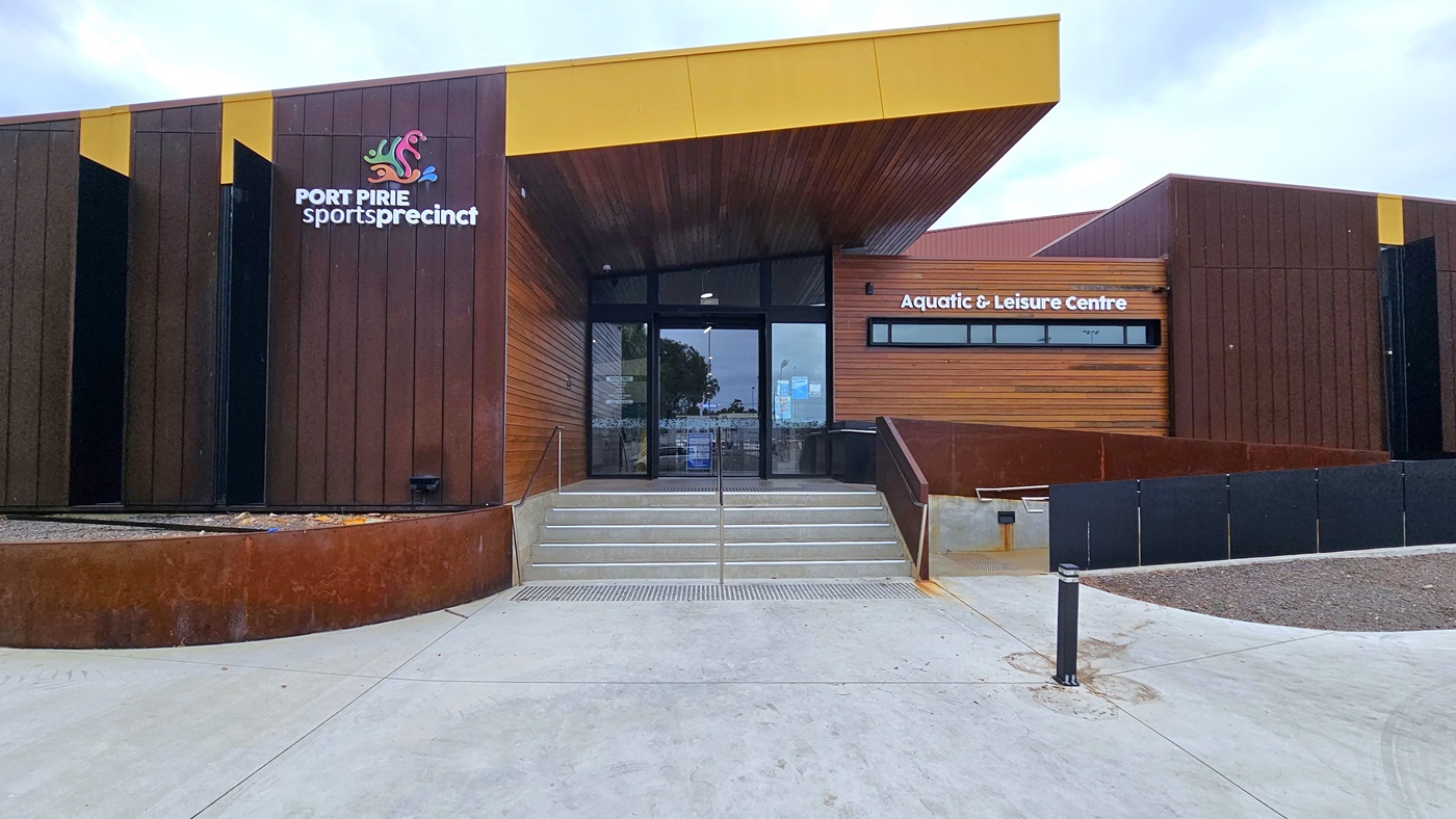 In the middle is a structure with modern architecture, displaying a sloping roofline, timber walls, and a warm brown timber ceiling. The facility signage is prominently placed at the front. Two sets of steps with handrails are in the middle, and a ramp entry with a railing is on the right. Truncated domes are installed before the steps. A black bollard and an empty concrete plant box are visible on the right.