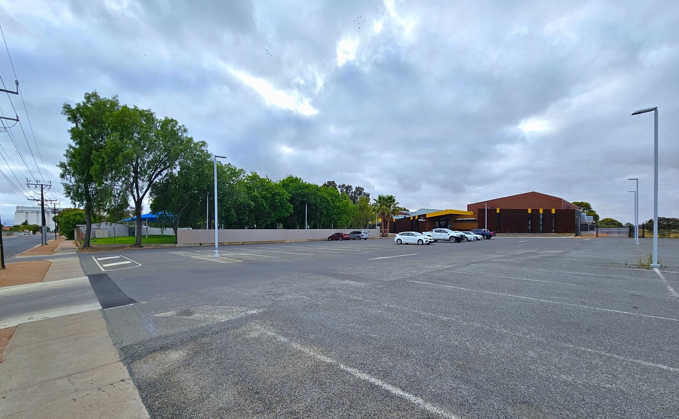 This is the expansive concrete car park adjacent to the Port Pirie Aquatic and Leisure Centre facility. The parking area is marked with white lines, and tall light posts are strategically positioned. In the middle, a few cars are parked. On the left is the main road, and further ahead, tall trees border the road and the grassy area.