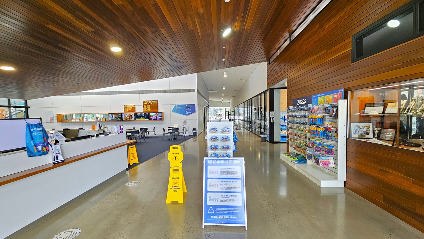 Entering, the reception is on the left with a glass display of achievements. Goggles and swimming accessories are on a rack to the right. A banner and safety signs are in the middle. Movable tables and chairs are on the left, flatscreen TVs are on the wall along with advert posters. The sloping timber ceiling has circular lights, and the floor is polished concrete.