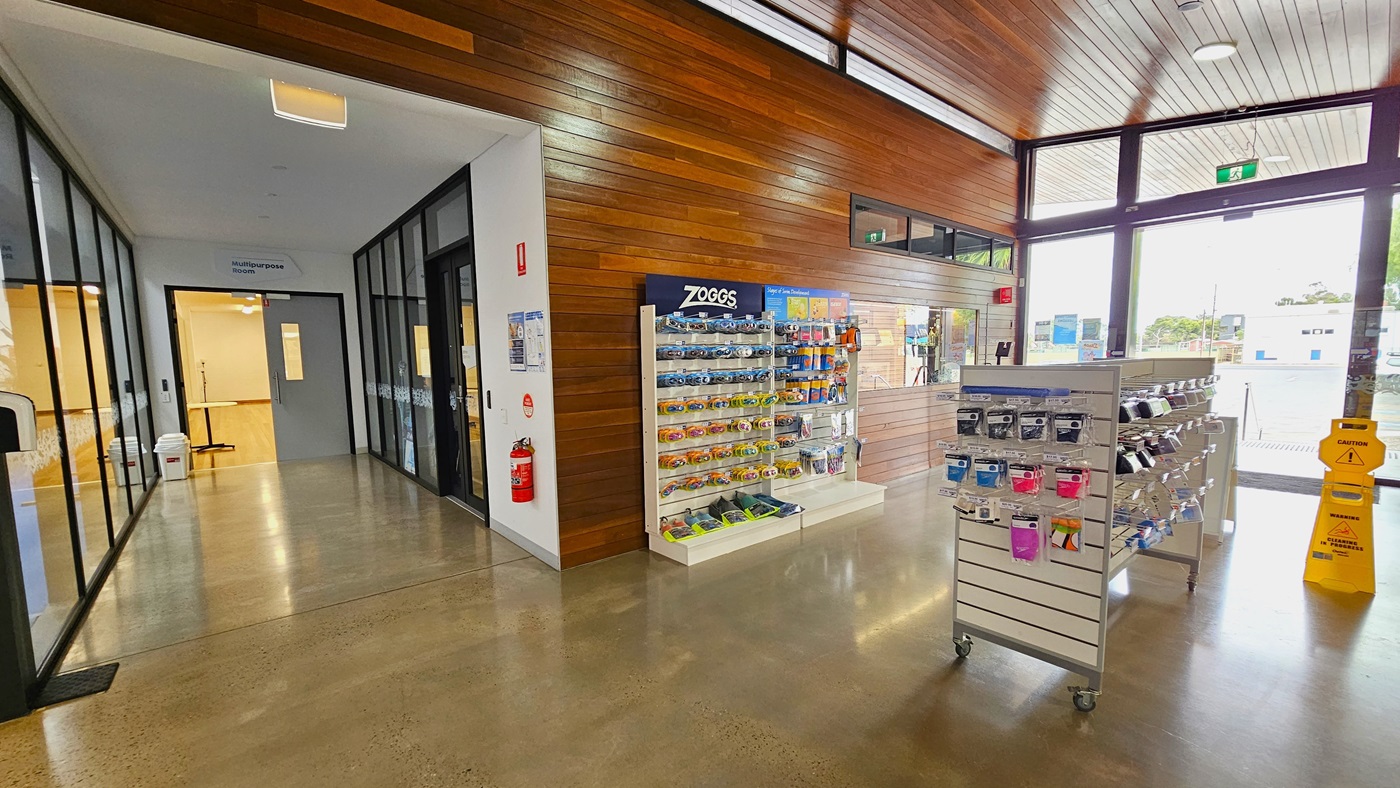 On the right is the glass door entry. A rack displaying goggles and swimming accessories is placed on the left, along with safety signs. To the left is a corridor that leads to the meeting rooms enclosed by floor-to-ceiling glass walls, and a manual door that opens inward. A rubbish bin is placed near the door. The floor has grey and cream tiles, and the ceiling is equipped with timber designs.