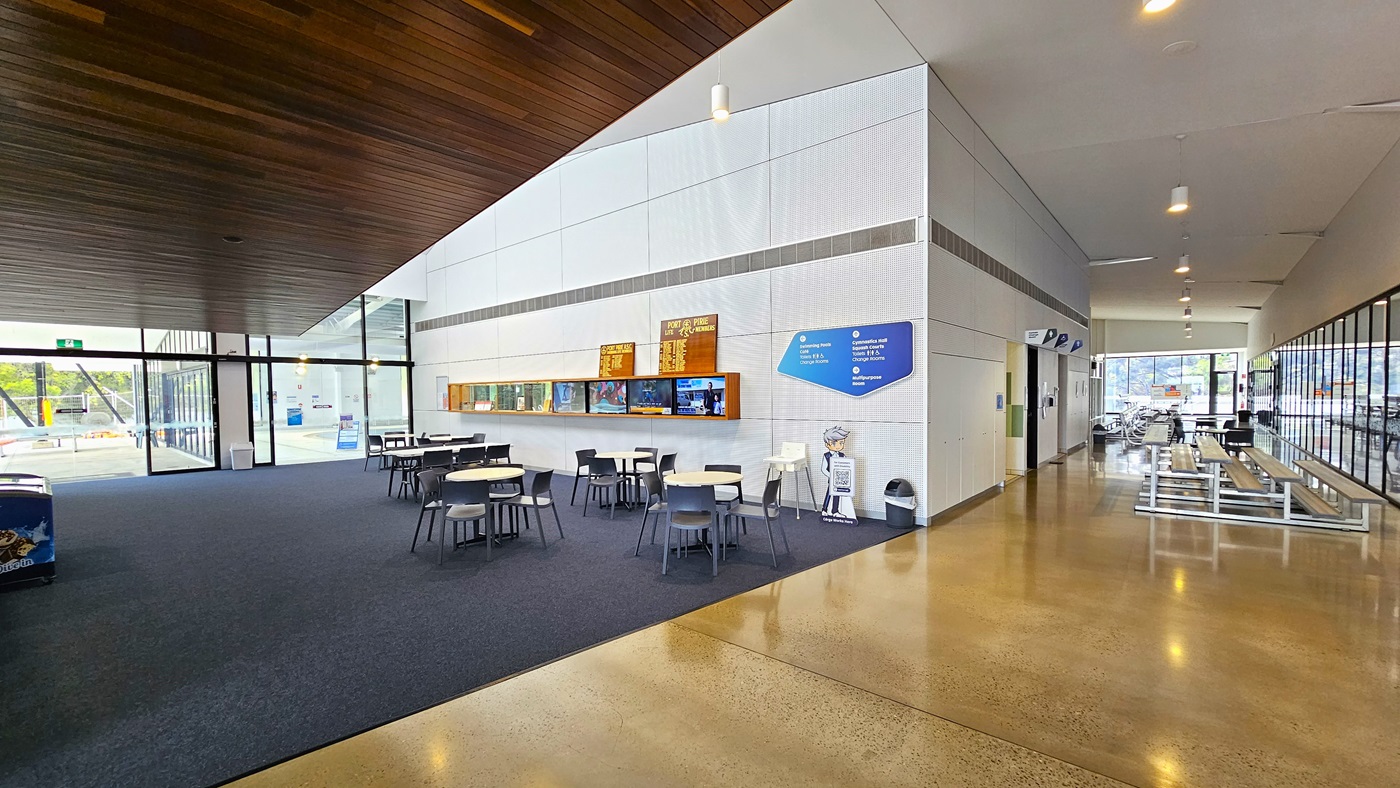 On the left is an ice cream fridge. Movable tables and chairs are arranged on the sides and near the white wall. Flat-screen TVs are mounted on the wall, along with adverts and facility achievements. A glass door is also positioned to the left. To the right is a corridor that leads to the accessible bathrooms and aquatic facilities. Tiered seating is available near the glass walls. The sloping ceiling features timber, and the floor is polished concrete and carpet.