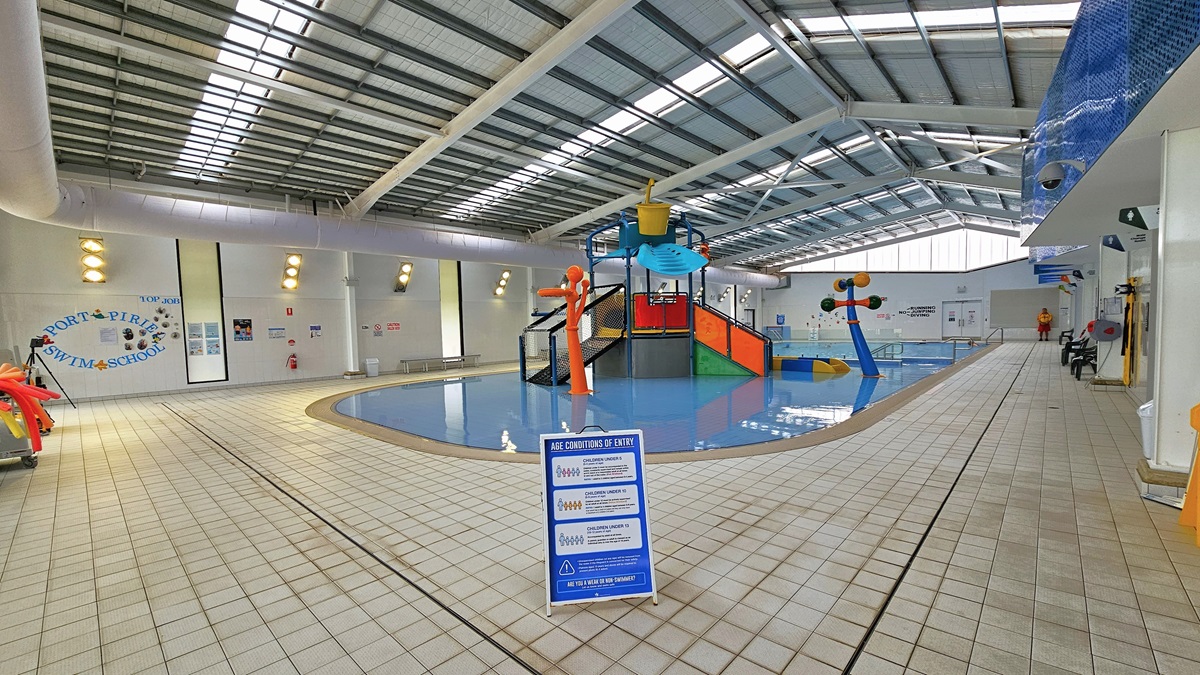 The entrance to the indoor Aquatic facility is through automatic glass sliding doors with posters. To the left, there's a poster and a white rubbish bin. Inside, to the right is the splash zone area with colorful slides and water features. To the left is a floor-to-ceiling glass wall with a view of the outdoor pool. A few free-standing banners are present, and the ceiling features exposed metal beams, insulated foams, and natural skylights.