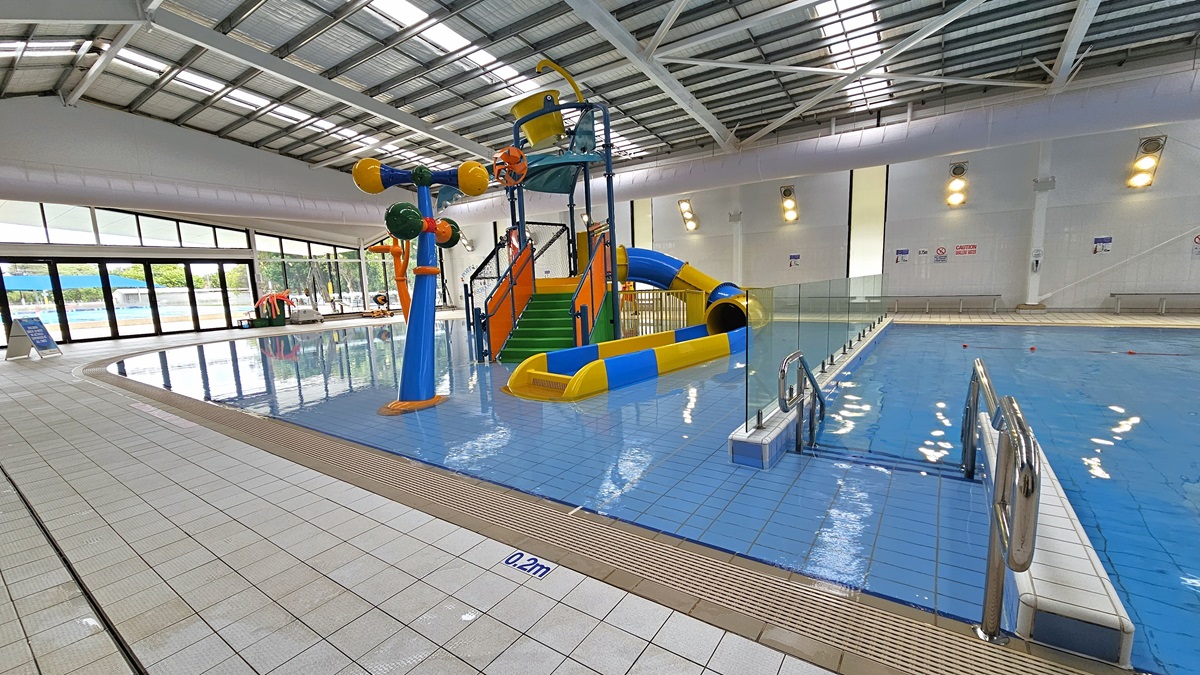 In the middle is a pool with colourful slides and water features. At the top of the slide is a yellow pail that pours water, along with orange and blue structures that spout water when in use. Accessible bathrooms are in the middle right, along with rows of circular lights. A low glass partition divides the splash zone from the bigger pool, which is equipped with a ramp entrance with handrails. The ceiling has exposed metal beams, insulated foams, and natural skylights.