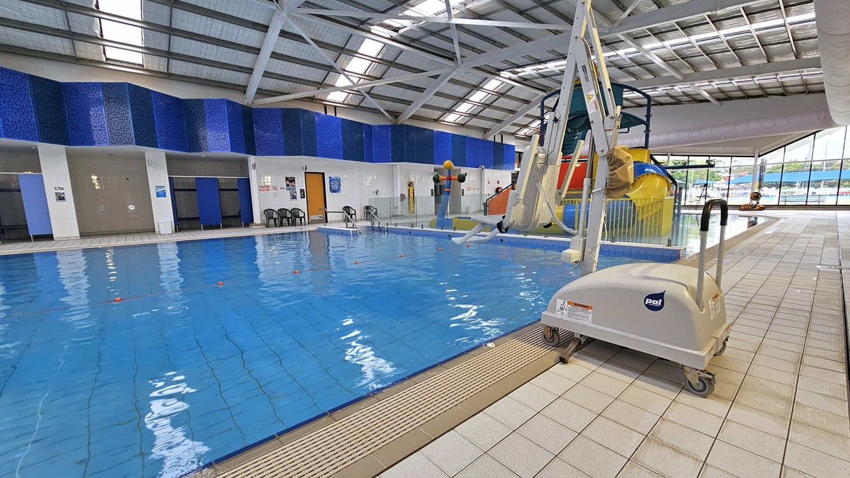 In the middle is the 15m indoor pool. A hoist is positioned on the right, near the pool overflow grid. Further right is the Splash Pool featuring a colourful slide with water features. On the opposite side of the pool are the accessible bathroom and change rooms. The walls on this side of the pool have a vibrant blue lining. The ceiling has exposed metal beams, insulated foams, and natural skylights.