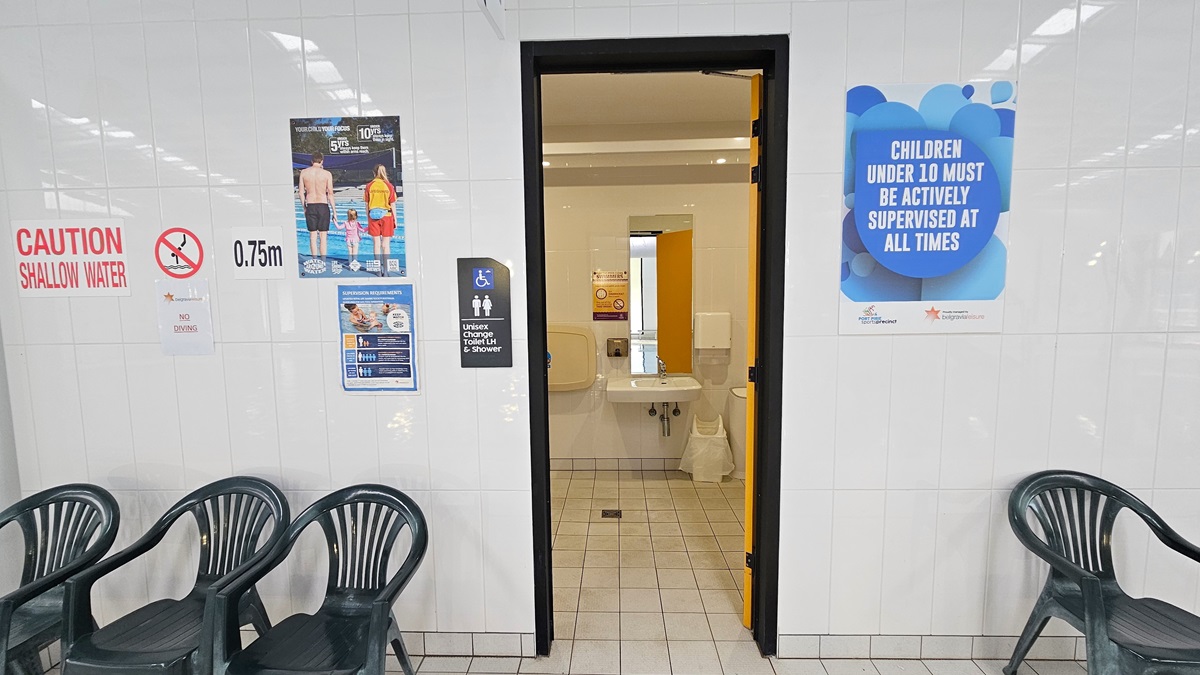The All-Gender Change Toilet and Shower features an inward-opening door. The left wall displays a gender indication sign and reminder posters. Three plastic chairs are aligned on the left, and another one is on the right of the door. The bathroom is adorned with white-tiled walls and floors. Inside, a wash basin, mirror, and rubbish bin are placed opposite the door.