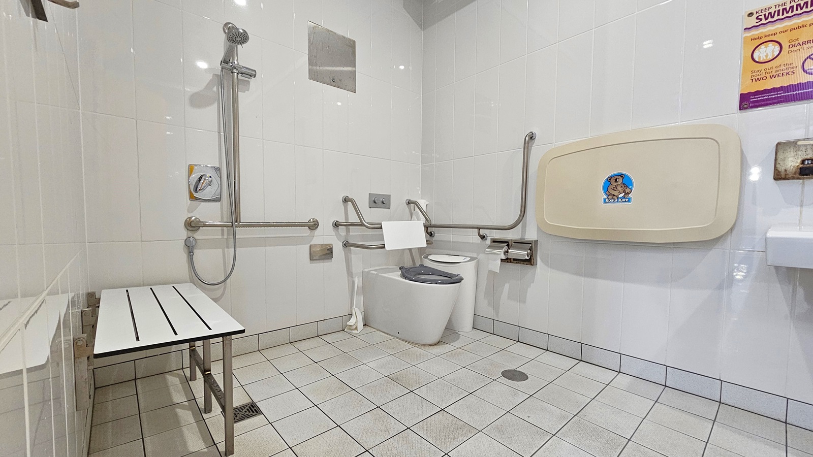 Inside the accessible bathroom. A toilet bowl with handrails is positioned in the right corner. Adjacent to it are the shower, tissue dispenser, and a rubbish bin. On the left, there is a bench mounted to the wall. To the right, a folded change table and wash basin are placed. The bathroom features all-white tiled walls and floor. A poster reminder is affixed to the right wall.