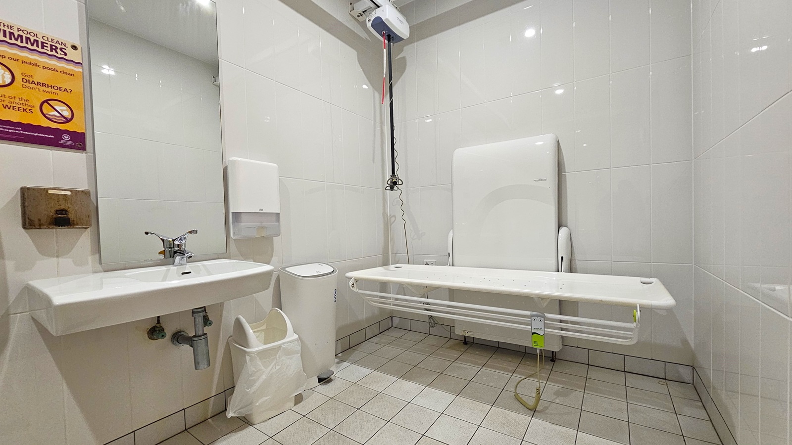 Inside the accessible bathroom. On the left, there is a wash basin, a mirror, and a tissue dispenser mounted to the wall. Adjacent are two rubbish bins. On the right is an adult-size changing table equipped with a hoist. The bathroom features all-white tiled walls and floor. A poster reminder is affixed to the right wall, and below it is a rusty metal structure.