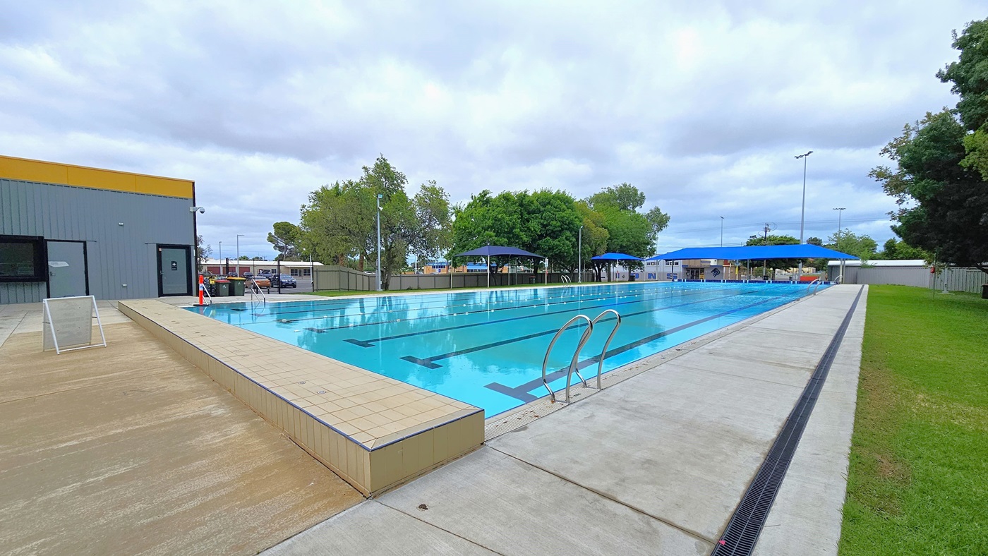In the middle is the 7-lane, 50-metre swimming pool equipped with handrails on each corner. An orange bollard is positioned at the corner. The pool coping is made of cream tiles, and a freestanding banner stands in the middle of the pool deck. On the left is a grassy area with shade cloths that extend towards the other side of the pool. On the right, there are chain fences and corrugated walls surrounding the area. Tall trees border the right side of the pool.