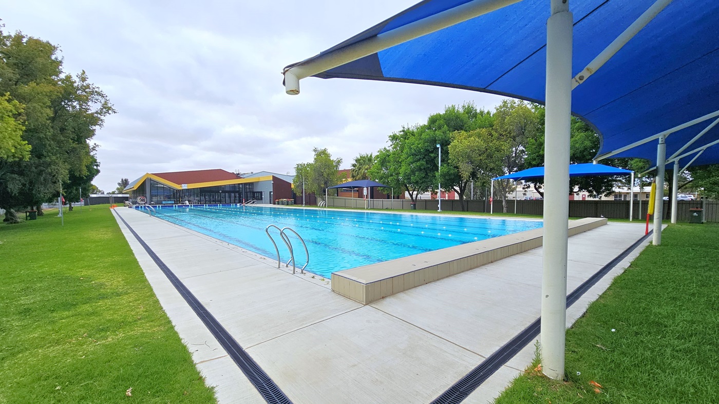 In the middle is the 7-lane, 50-metre swimming pool equipped with handrails on each corner. An orange bollard is positioned at the corner. The pool coping is made of cream tiles, and a freestanding banner stands in the middle of the pool deck. On the right is a grassy area with tall trees. On the left is a building with corrugated grey walls and a yellow roofline. Shade cloths are positioned at the far end.