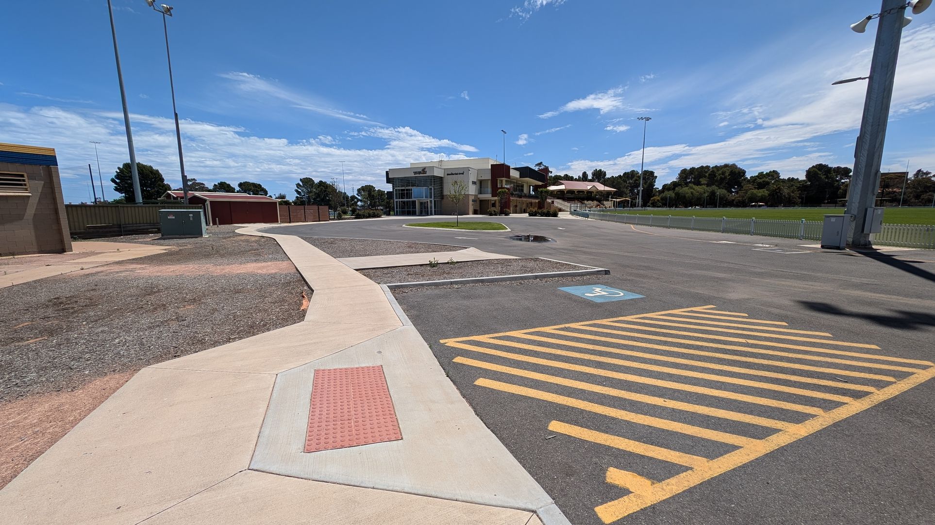 A parking area with two marked disabled parking spaces on the asphalt. Yellow diagonal lines separate the spaces. On the left, a low, beige building with blue trim and a covered walkway. In the background, tall light poles and a fenced area. On the right, a modern building with a curved facade and a small tree on a grassy patch. The ground is mostly paved, with a small puddle visible.