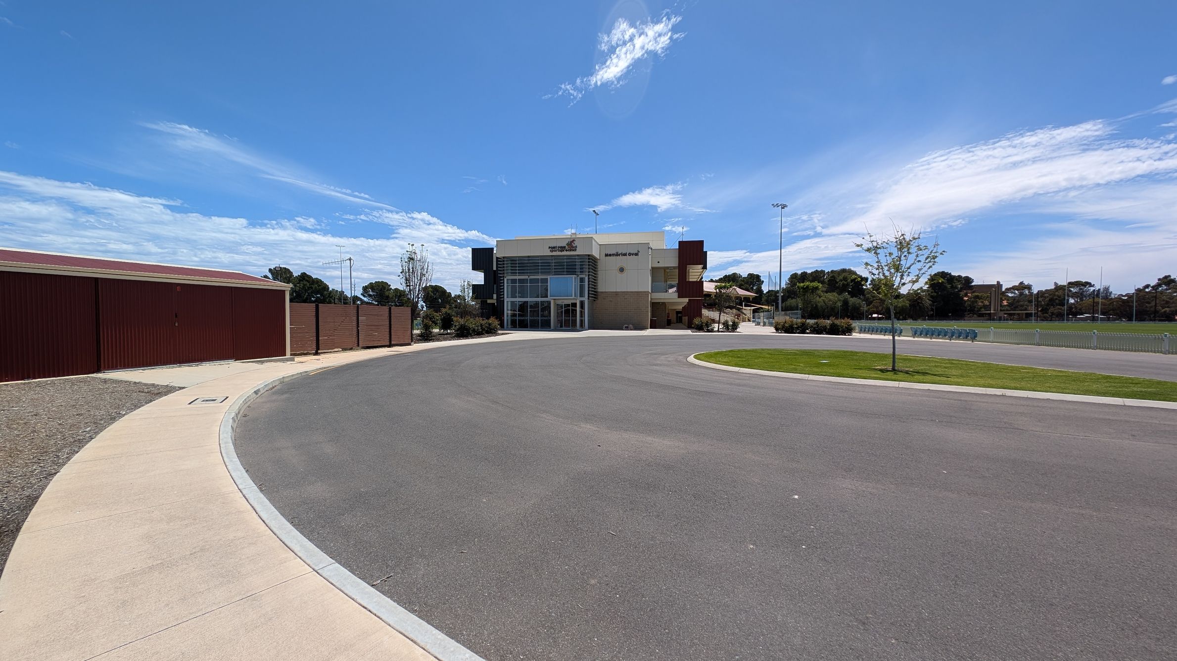 A modern building with large glass windows is centred in the background, labelled "Memorial Oval". To the left, a long, brown fence runs alongside a concrete path. On the right, a grassy area with a young tree is visible, adjacent to a paved area with a row of bollards. The foreground features a wide, empty asphalt road curving towards the building. Trees and a sports field with tall lights are in the distance.
