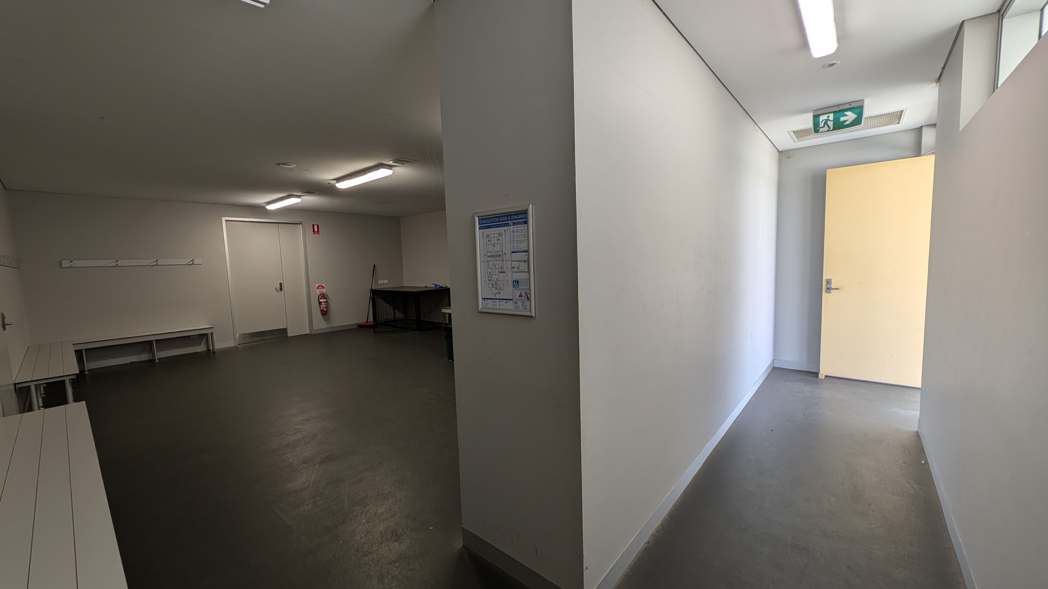 A corridor on the right with a light grey floor and white walls, leading to an open door. An emergency exit sign is above. On the left, a larger room with benches along the walls, a fire extinguisher, and a closed door. The ceiling has fluorescent lights. A wall with a diagram separates the two areas.