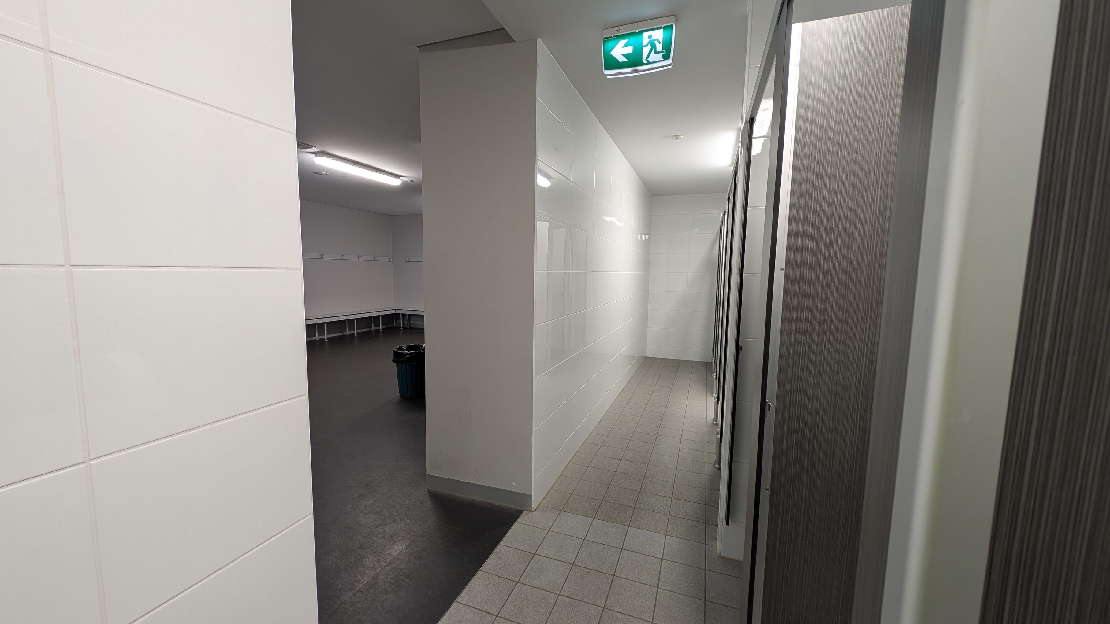 A corridor with white tiled walls and a green-tiled floor leads to a row of cubicles on the right. Above, an illuminated green exit sign with an arrow points left. On the left, an open area with dark flooring features benches along the walls and a rubbish bin. The ceiling is white with fluorescent lighting.