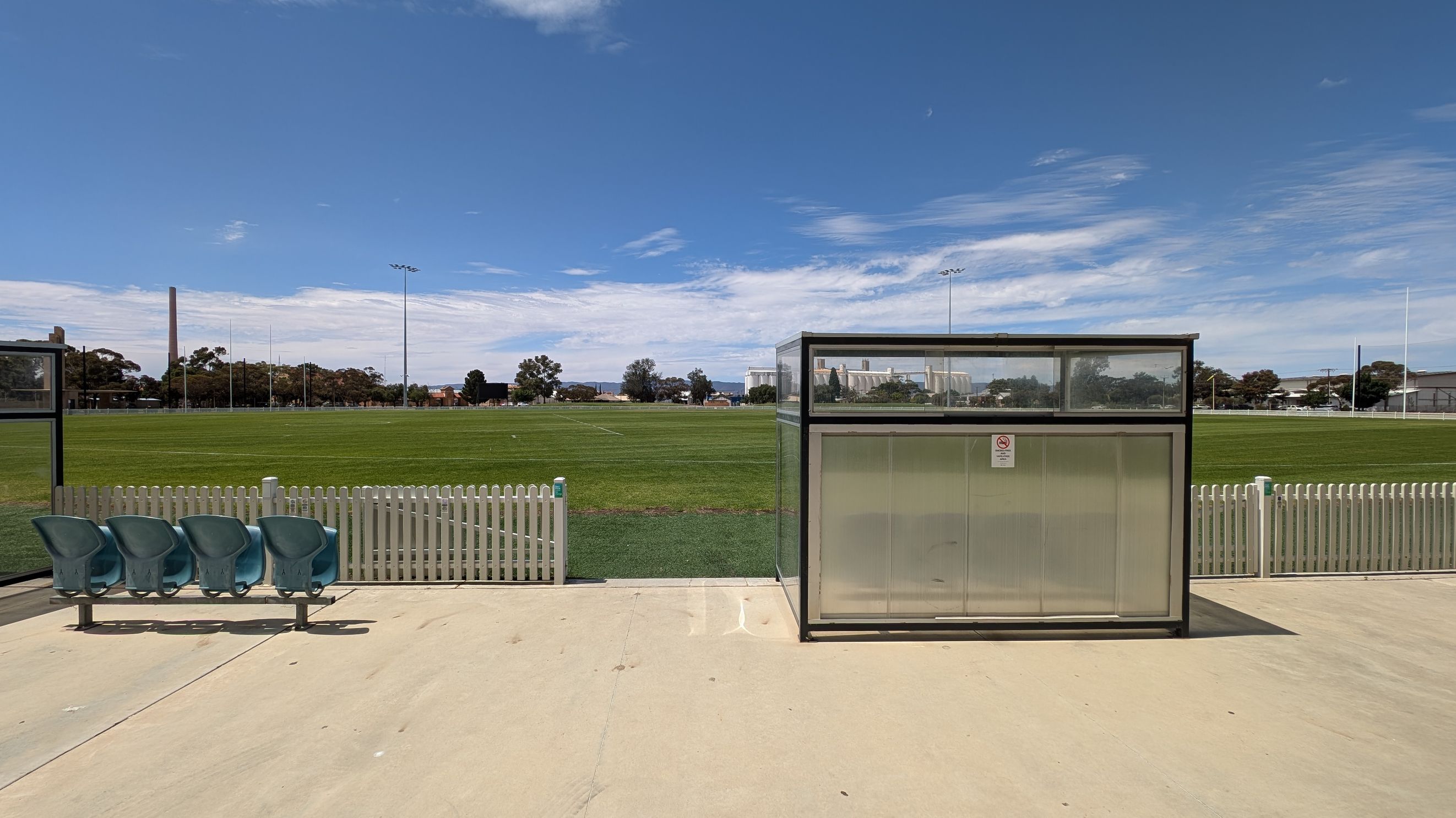 A sports field with a green grass pitch in the background, surrounded by a white picket fence. In the foreground, there is a small shelter with a transparent roof and walls on the right, and a row of four green plastic seats on the left. The ground is paved with light-coloured concrete. Tall light poles and trees are visible in the distance.