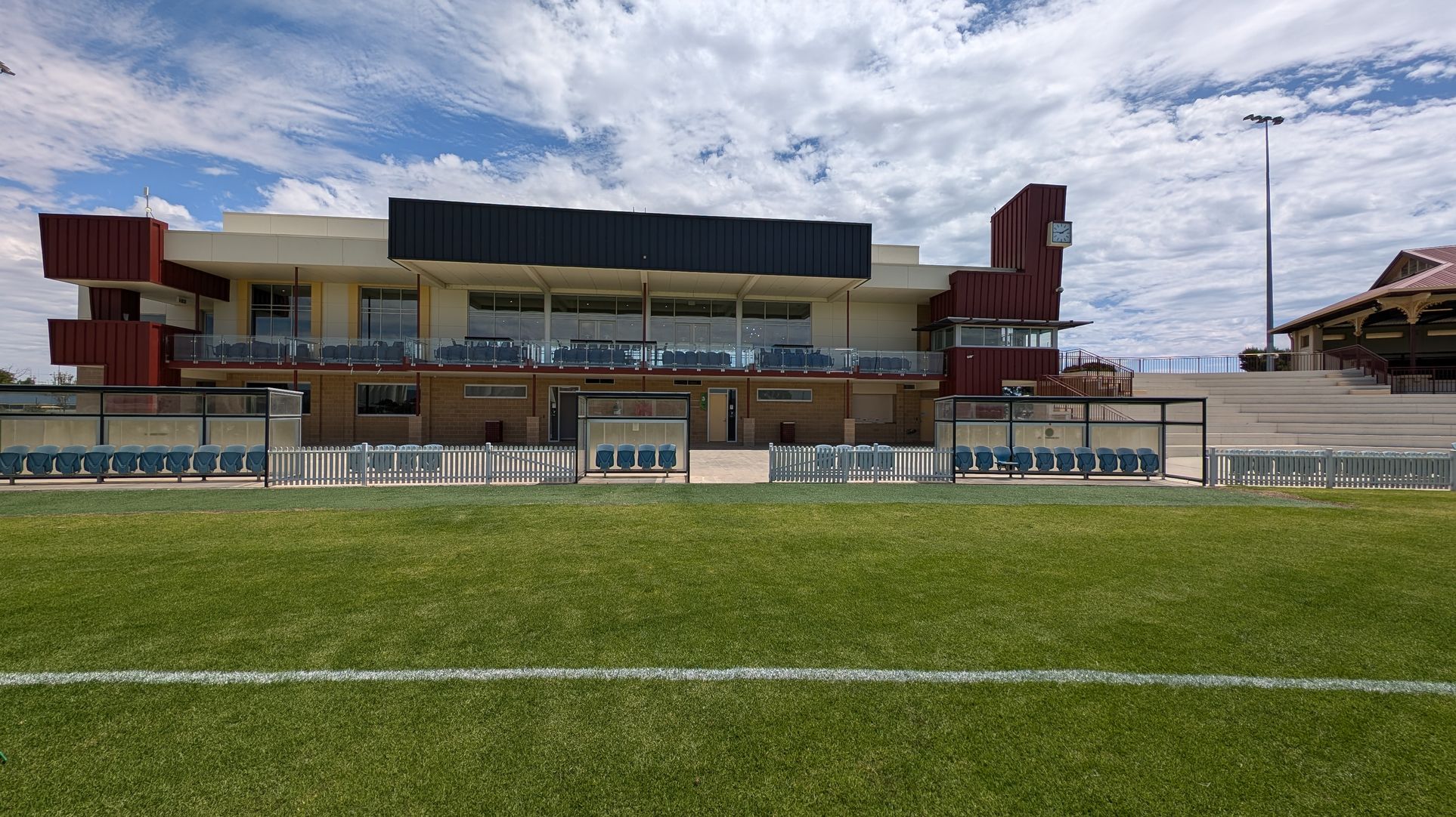 A sports stadium with a modern two-storey building in the background. The building features large glass windows and a mix of red and black panelling. In front of the building, there are two covered seating areas with blue chairs, separated by a white picket fence. To the right, there are concrete steps leading up to additional seating. The foreground shows a well-maintained grass field with a white line marking.