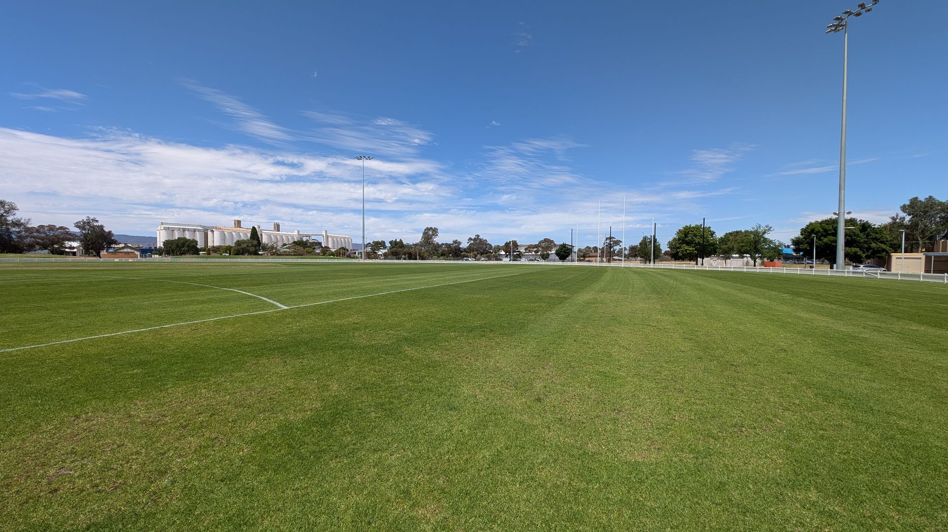 A large, well-maintained grassy sports field with white line markings. On the right, tall floodlights stand along the perimeter. In the background, there are several trees and a row of industrial buildings with cylindrical structures. The field is surrounded by a low white fence, and the sky is clear with some clouds.